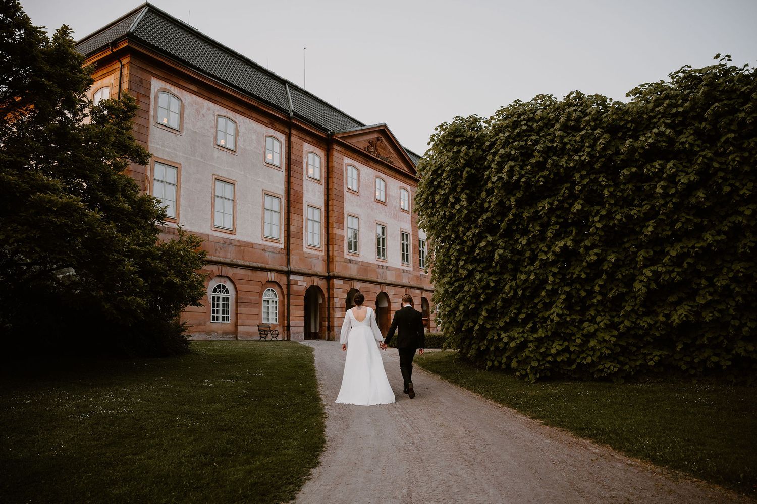 Two figures walk along a garden path outside a grand brick building with surrounding greenery and ivy.