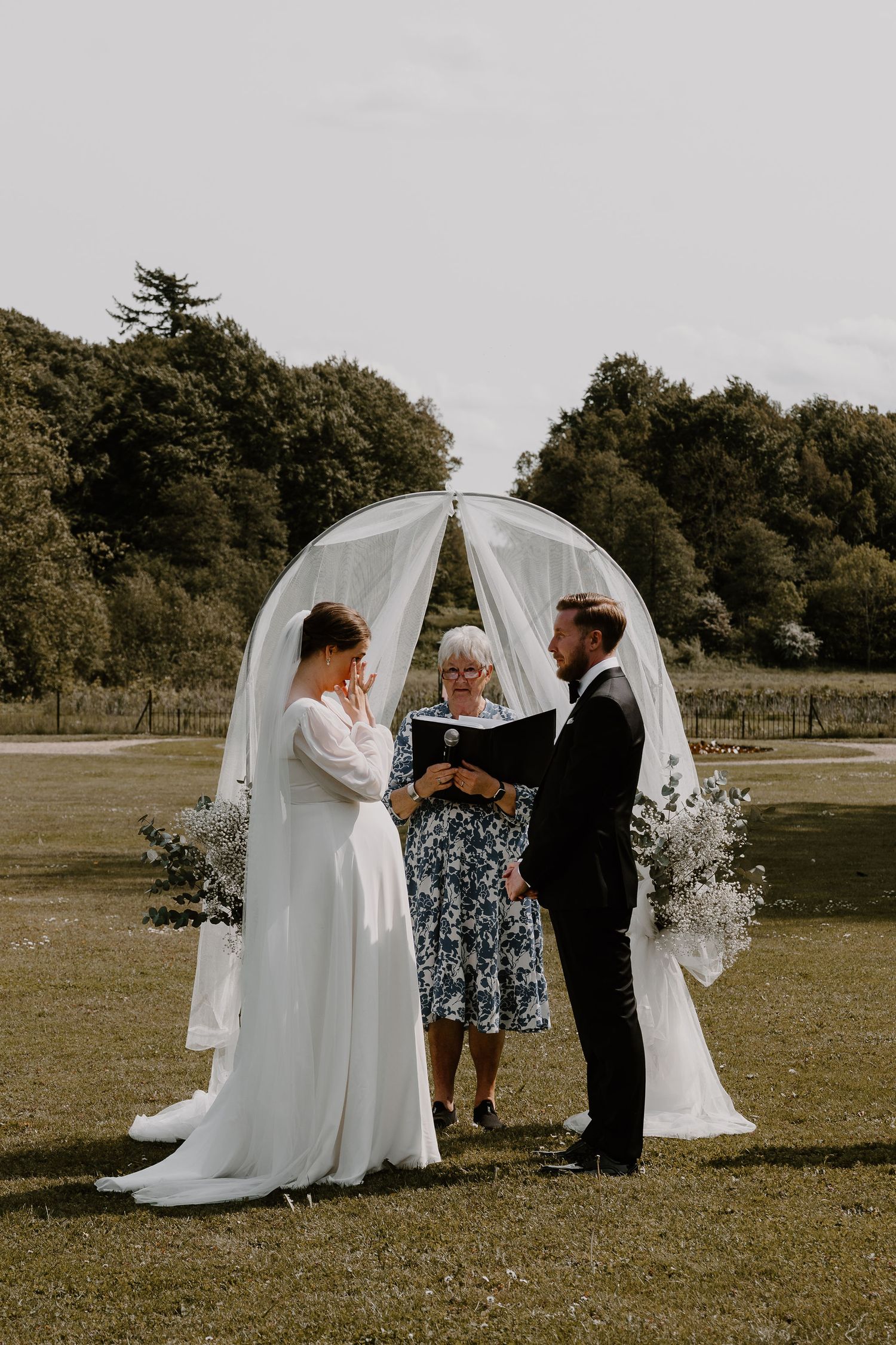 A couple shares an intimate moment during their outdoor wedding ceremony under a graceful white arch on a grassy field.