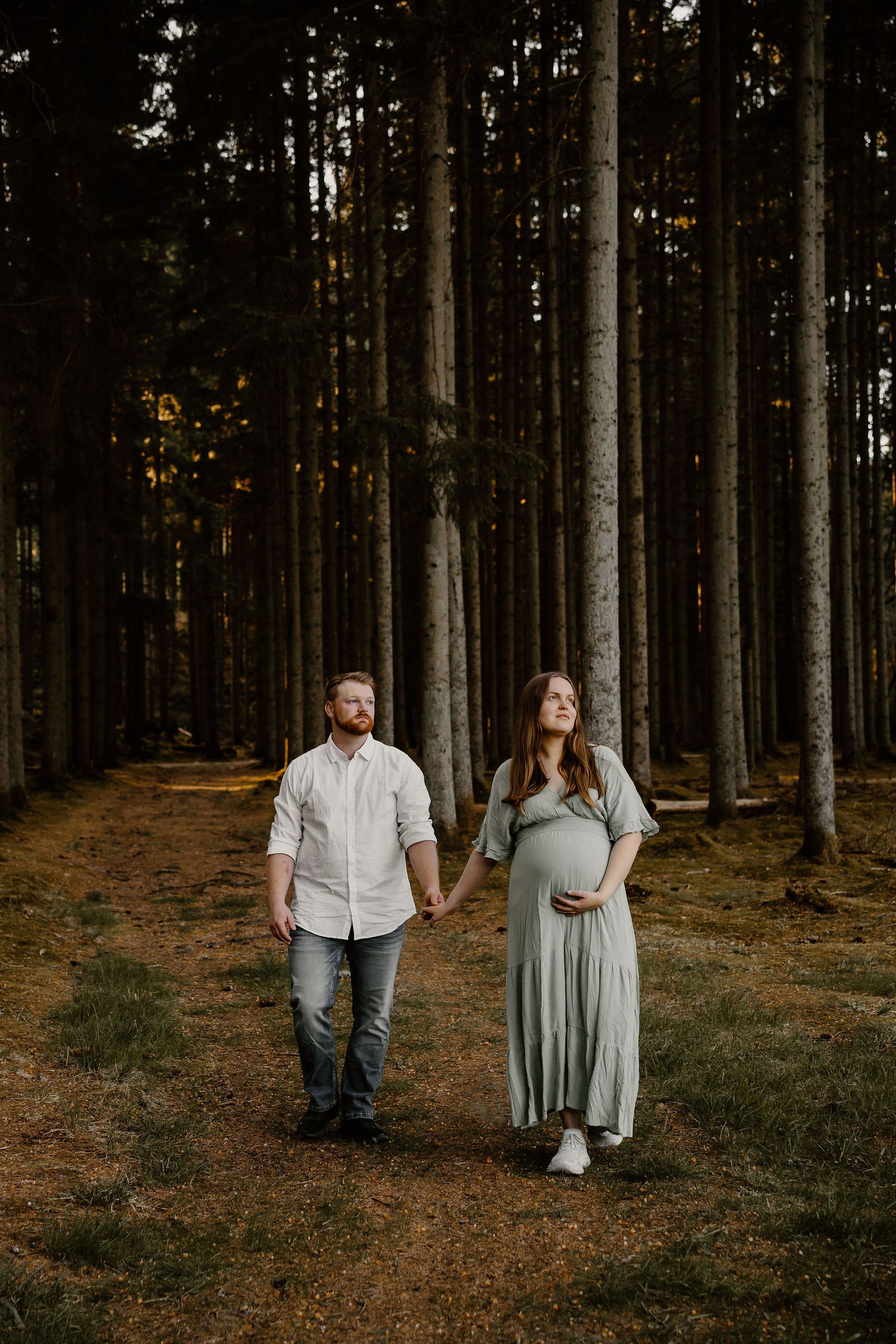 A couple stands together on a forest path surrounded by tall pine trees at sunset, with the woman wearing a flowing dress.