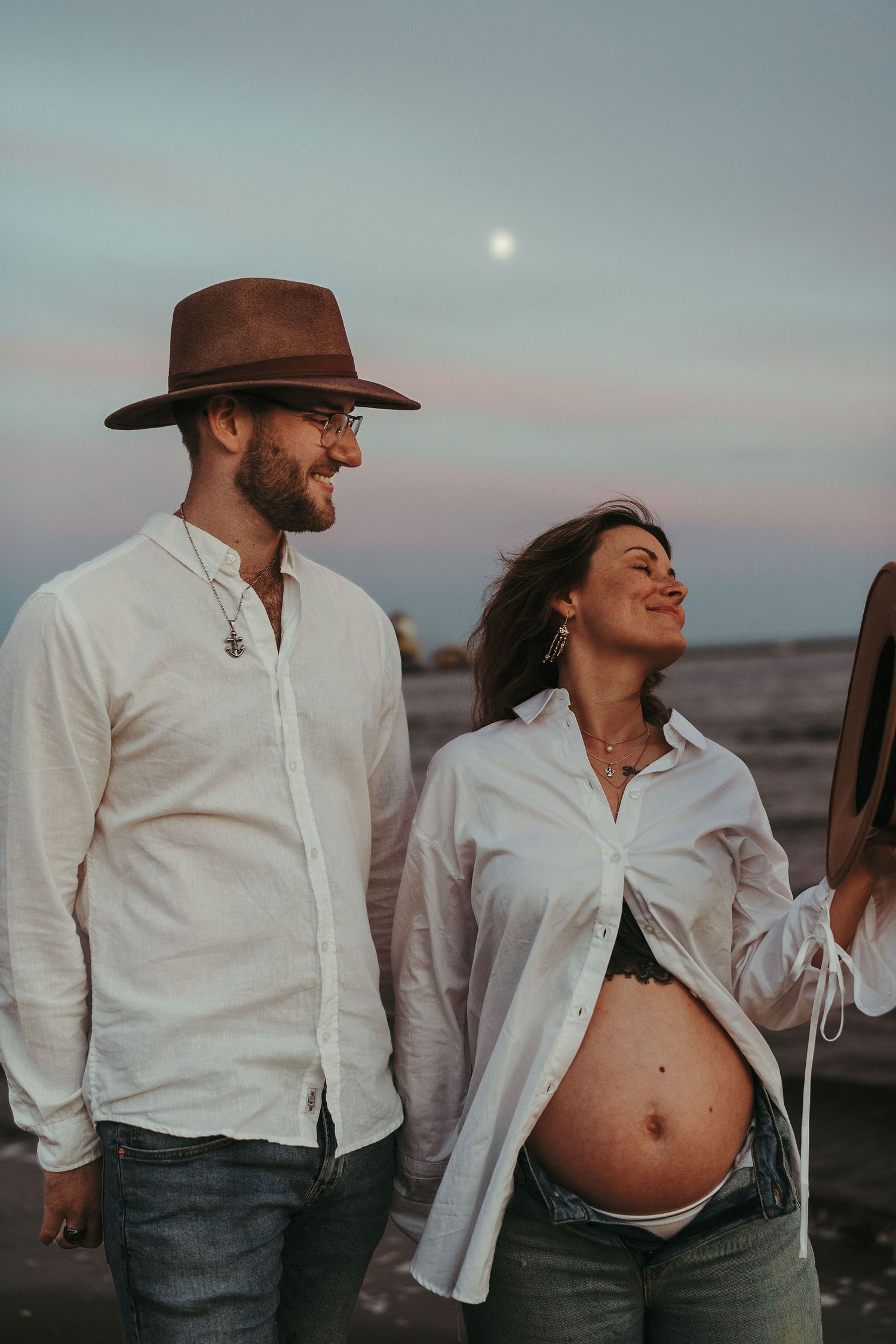 A couple in white shirts stands outdoors at dusk with a moon visible, showing off a pregnant belly.