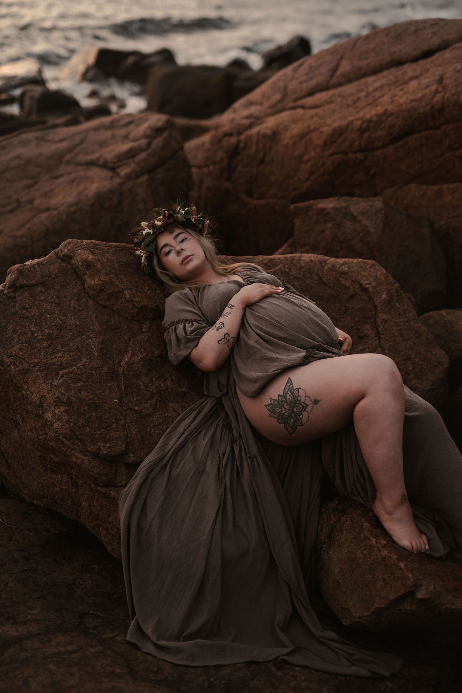 A figure in a brown flowing dress reclines on coastal rocks wearing a floral crown at sunset.