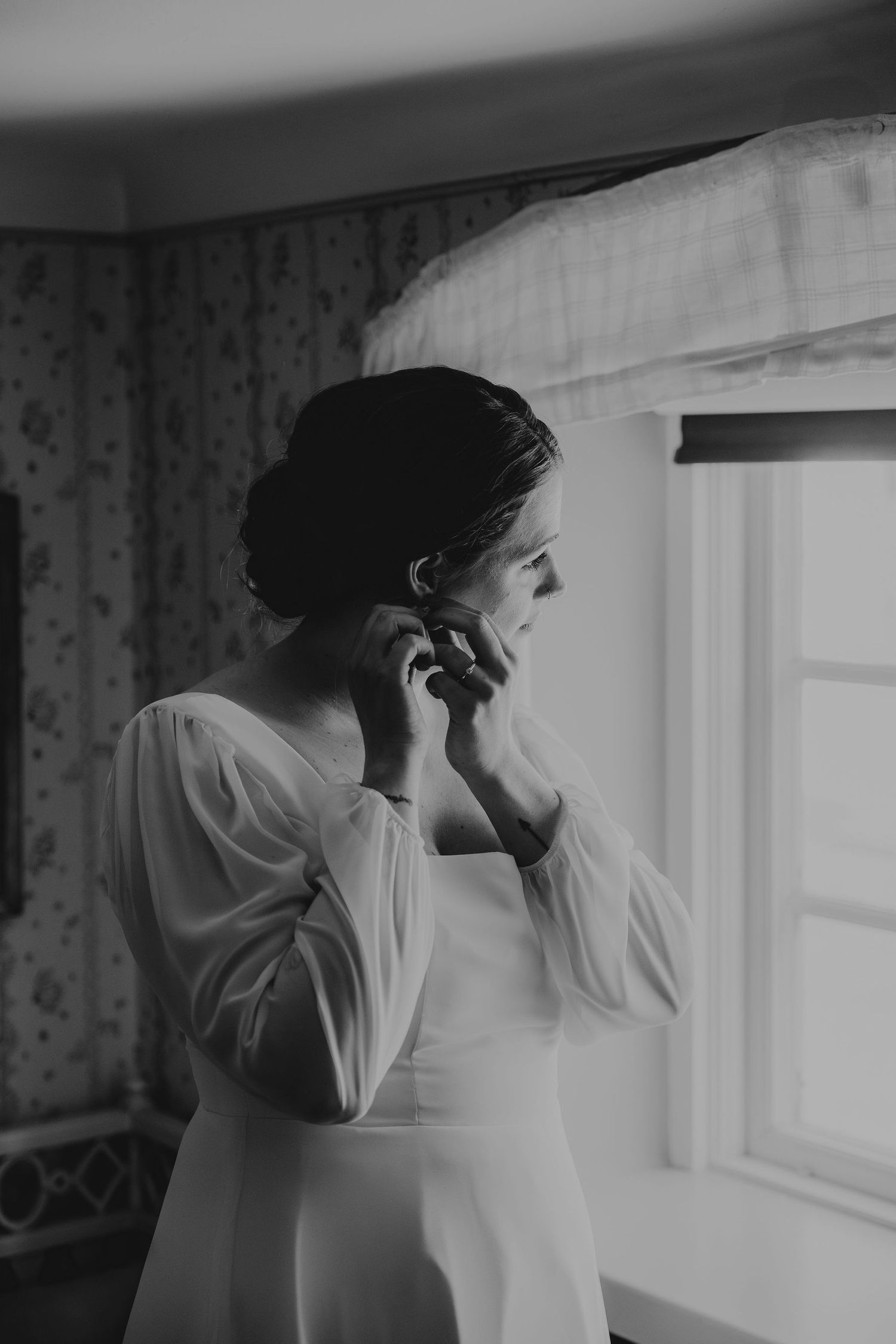 A silhouetted figure in a white dress stands by a window while talking on the phone in a black and white photograph.