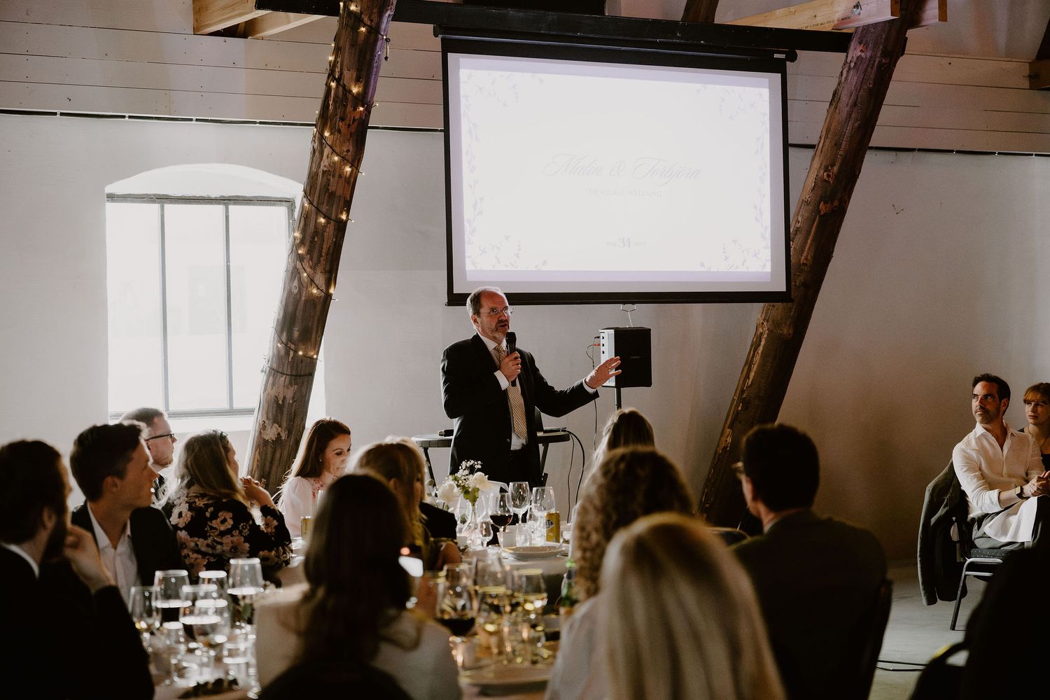 Indoor reception venue with exposed wooden beams and warm lighting creates an intimate dining atmosphere with multiple guests.