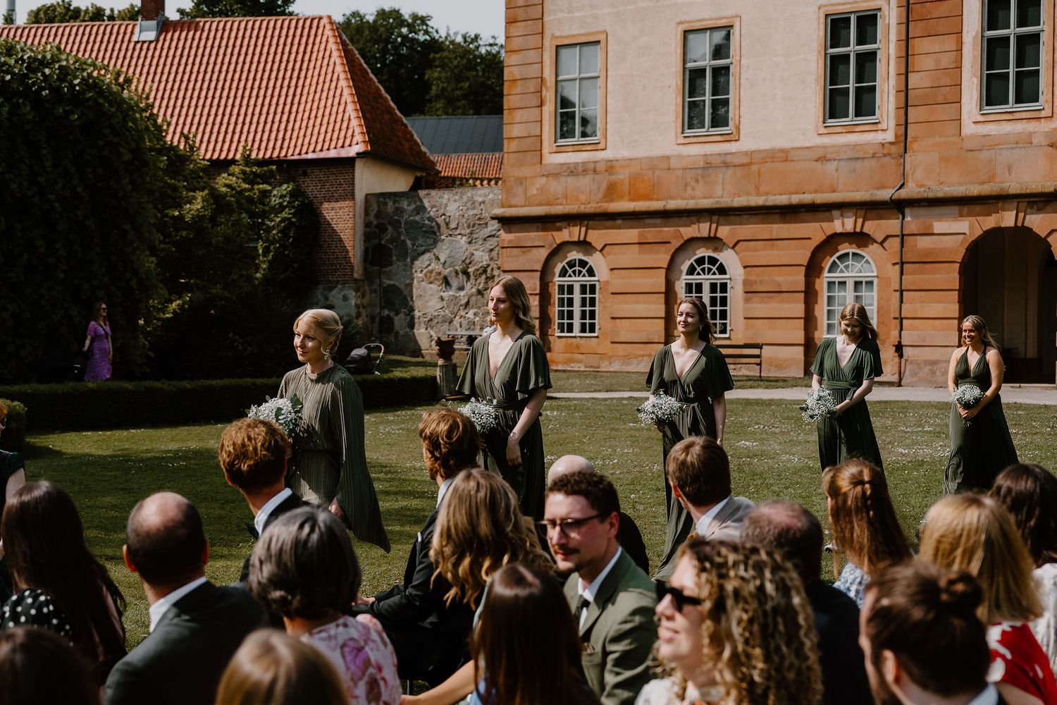 A summer wedding celebration takes place in the courtyard of a historic building with orange walls and arched windows.