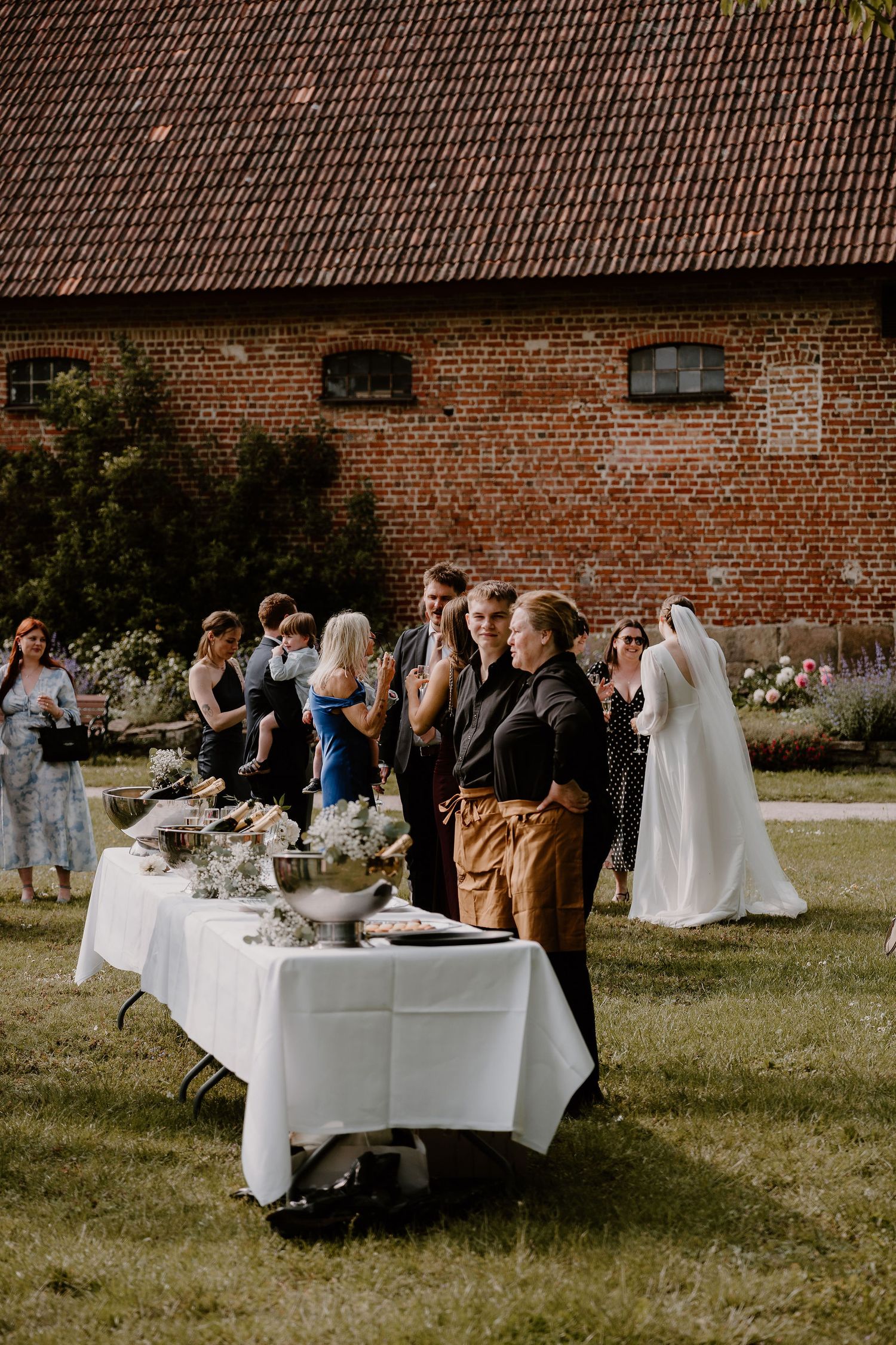 Wedding guests gather around elegant white-clothed tables set up outdoors against a rustic brick building backdrop.