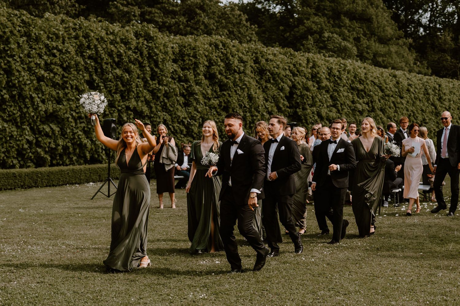 Wedding party walks across green lawn during outdoor ceremony with formal attire and bouquets.