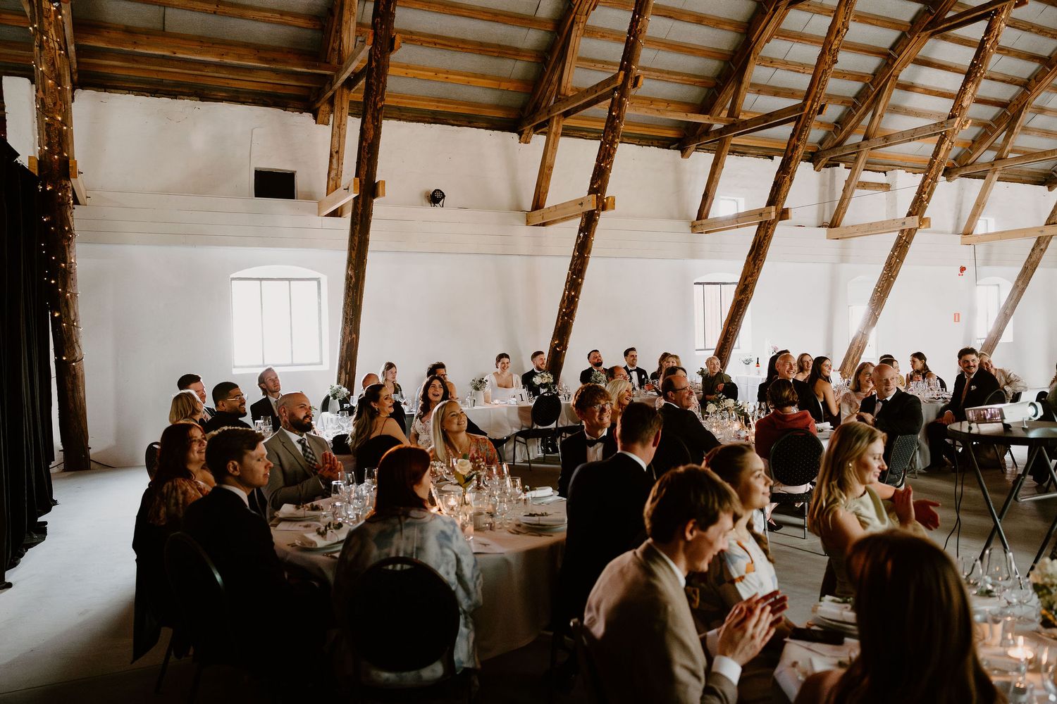 Wedding reception guests seated at round tables in a rustic white barn venue with exposed wooden beams and natural light.