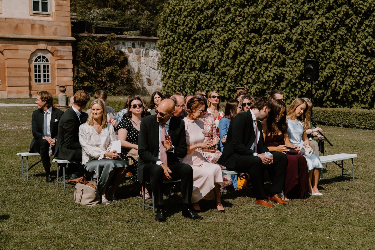 Wedding guests gather on benches in the sunny garden courtyard for an outdoor ceremony.