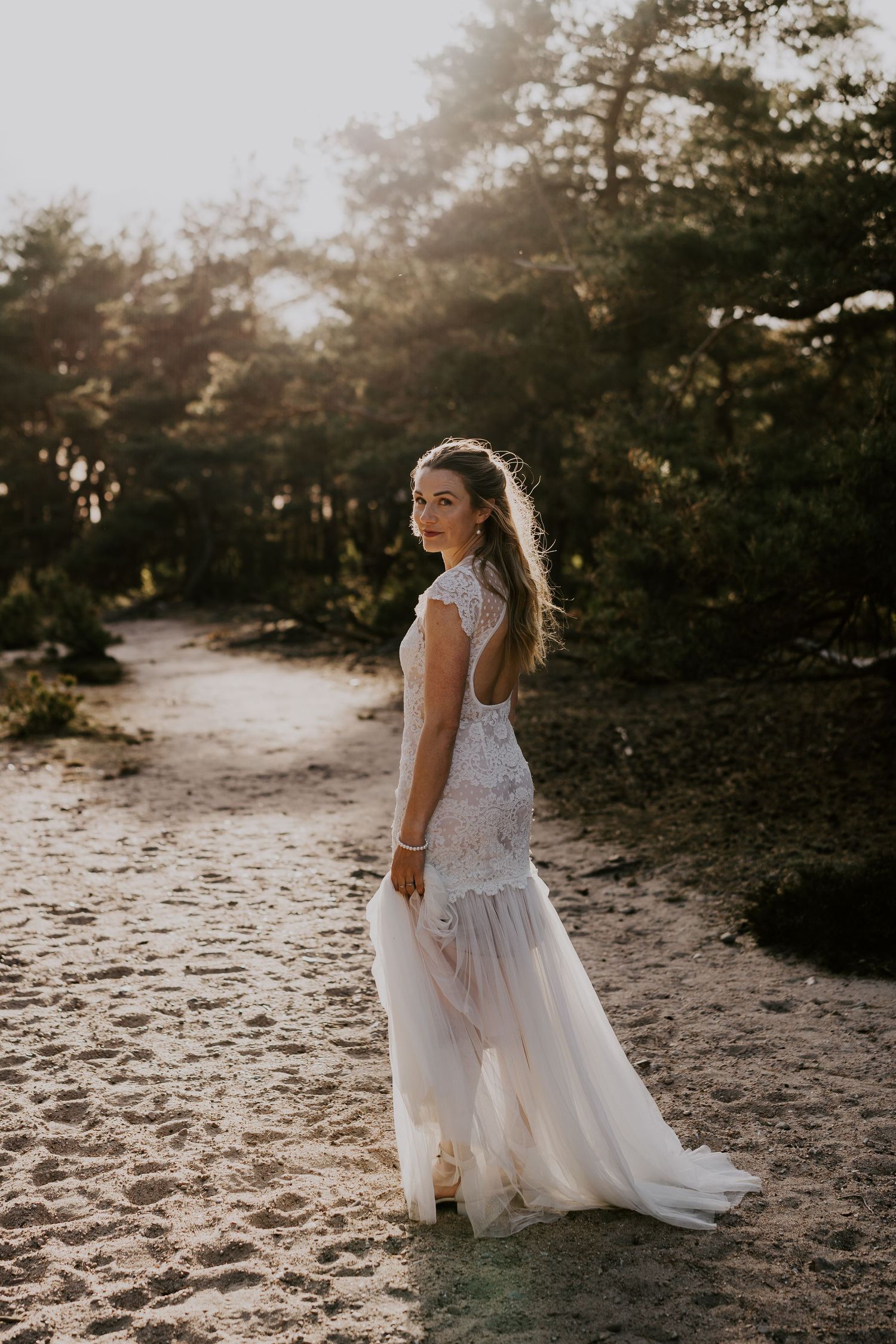 Beautiful lace wedding dress shot with golden sunlight streaming through pine trees on sandy pathway.