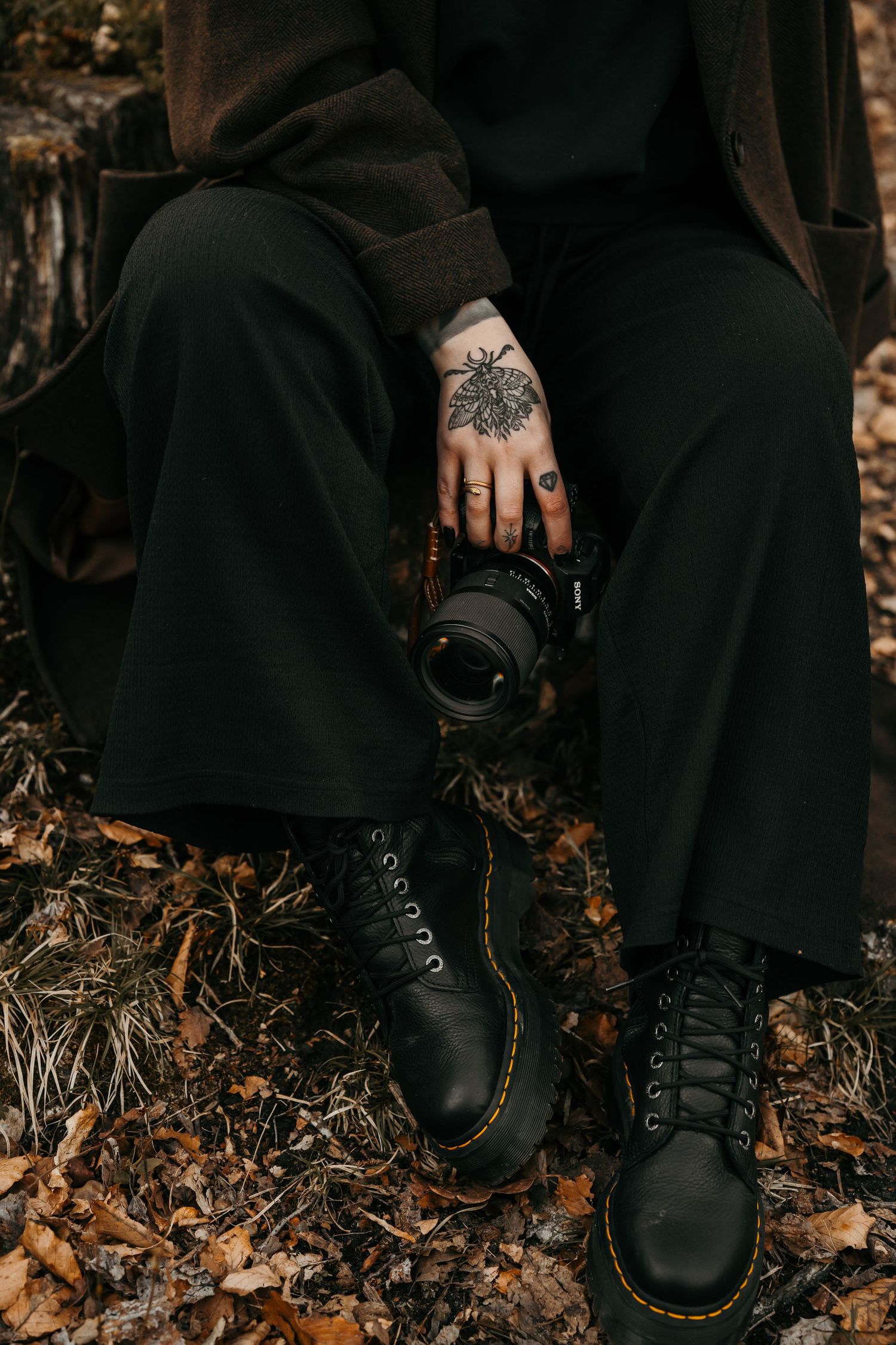 Person in black boots and clothing holding a camera displays intricate mandala hand tattoo among autumn leaves.