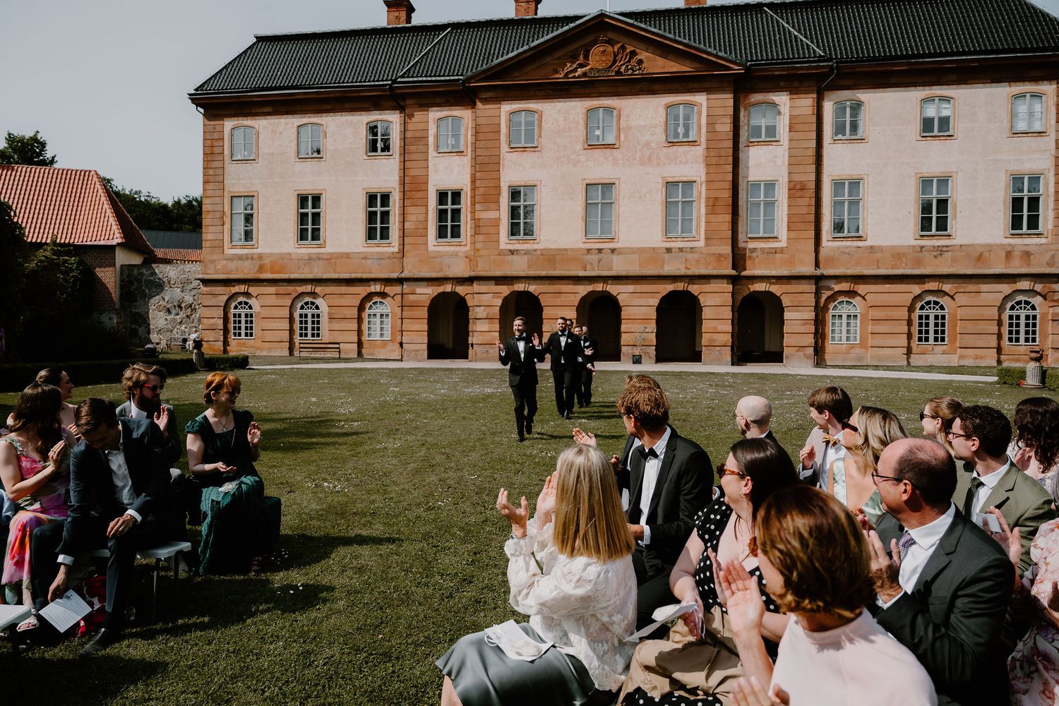 A wedding ceremony taking place on a lawn outside a grand historic mansion with pink and beige facade.