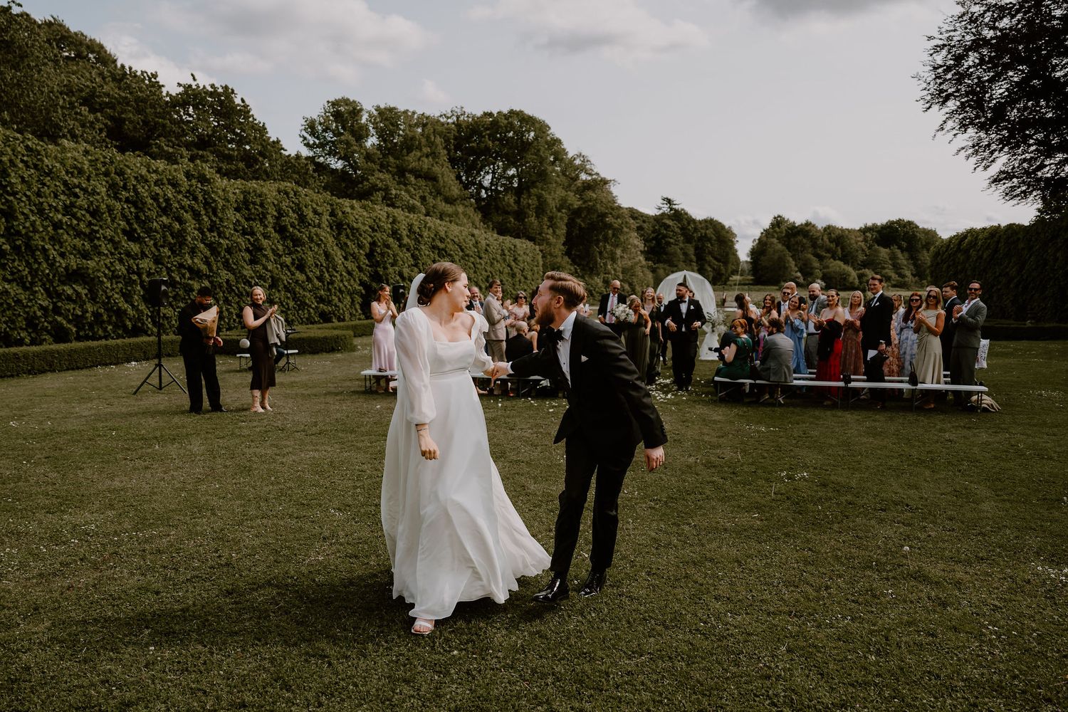 A garden wedding celebration on a manicured lawn with guests gathered around watching the newlyweds.