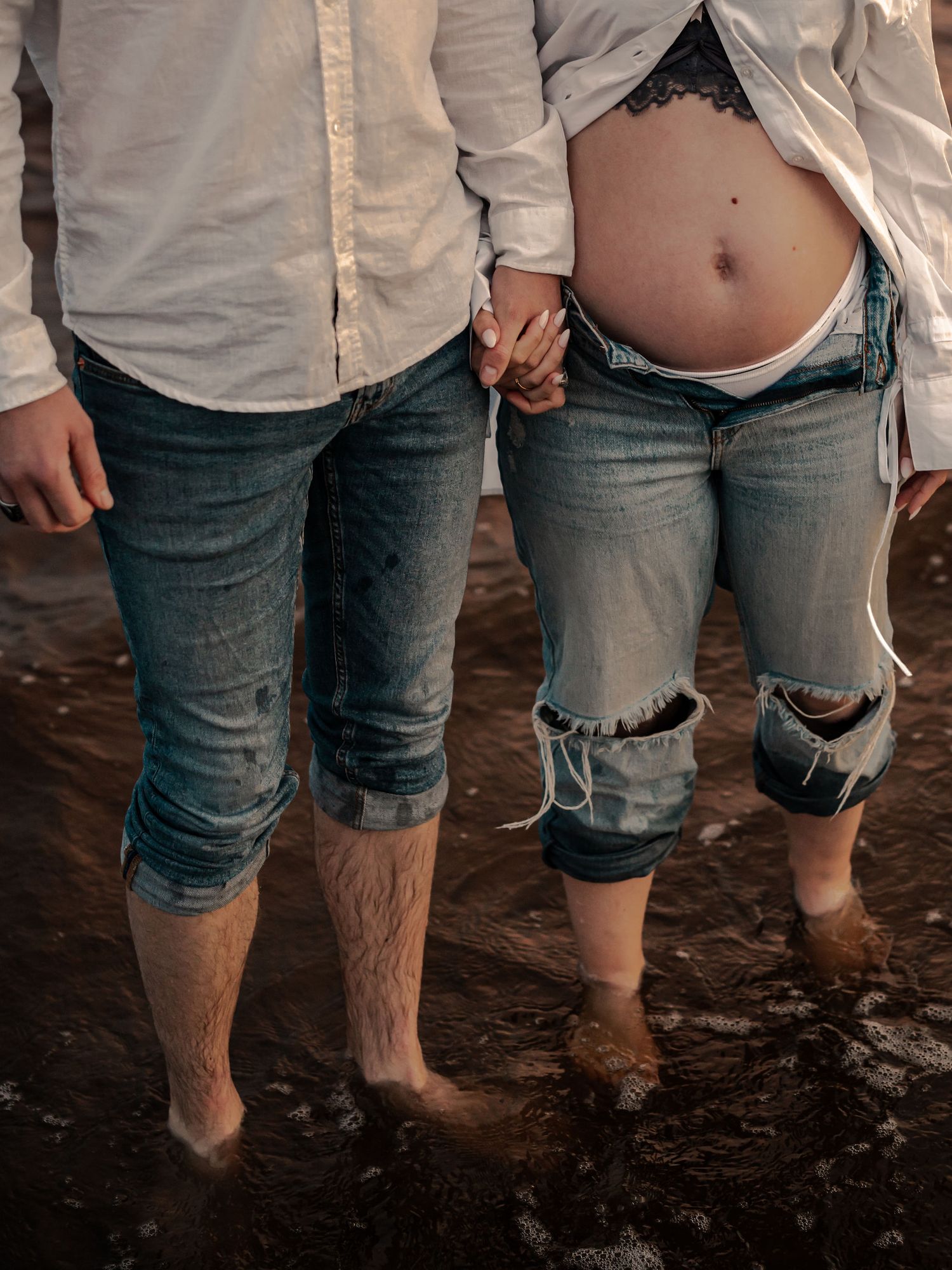 Couple holding hands in ripped jeans standing barefoot in shallow water on a beach.