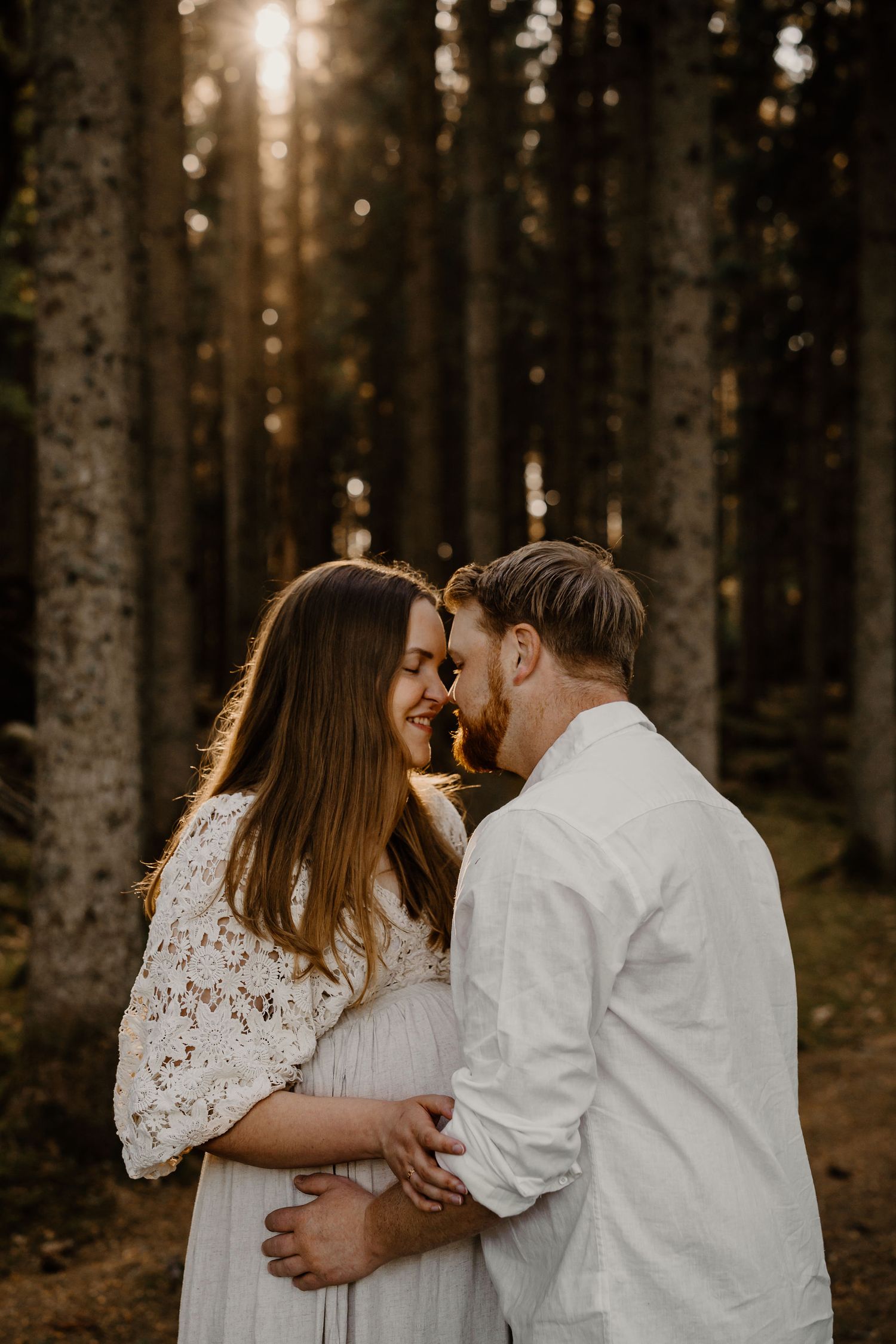 A couple in white outfits embrace in a sunlit forest during a romantic maternity photoshoot at golden hour.