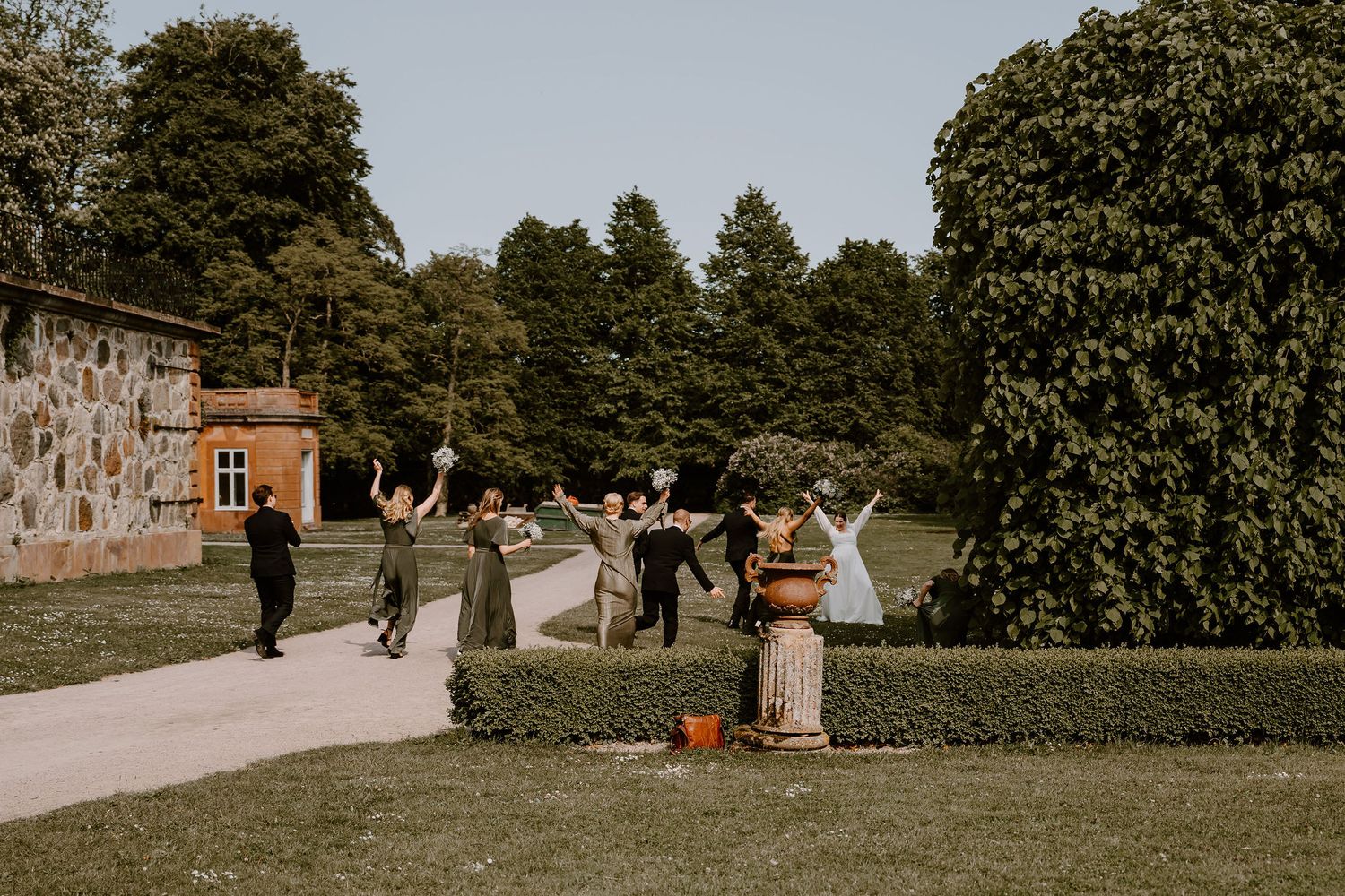 Wedding guests gather on a scenic garden path beside stone walls and lush greenery on a bright afternoon.