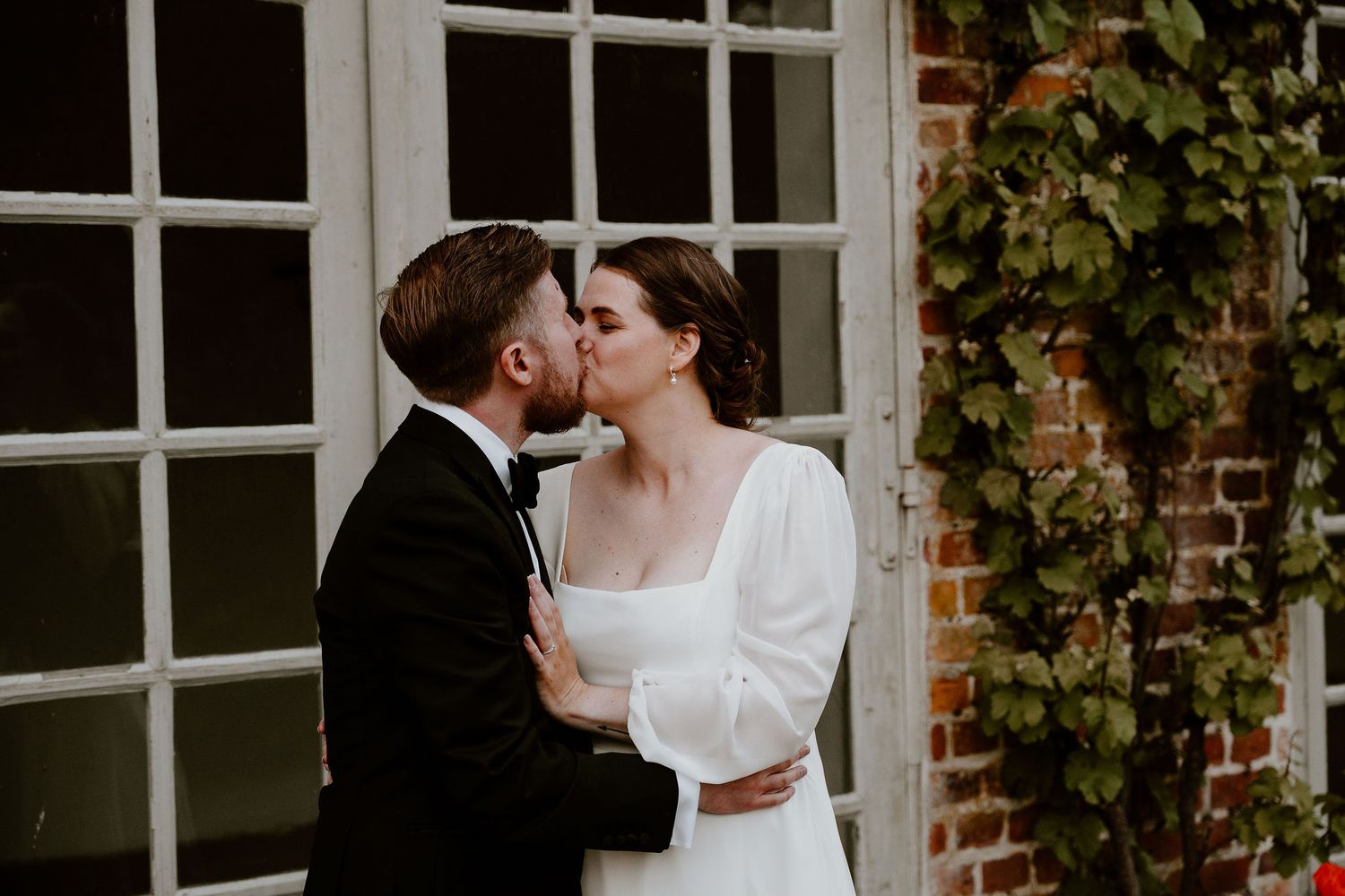 Newlywed couple shares intimate outdoor moments by white french doors and brick walls with climbing ivy.