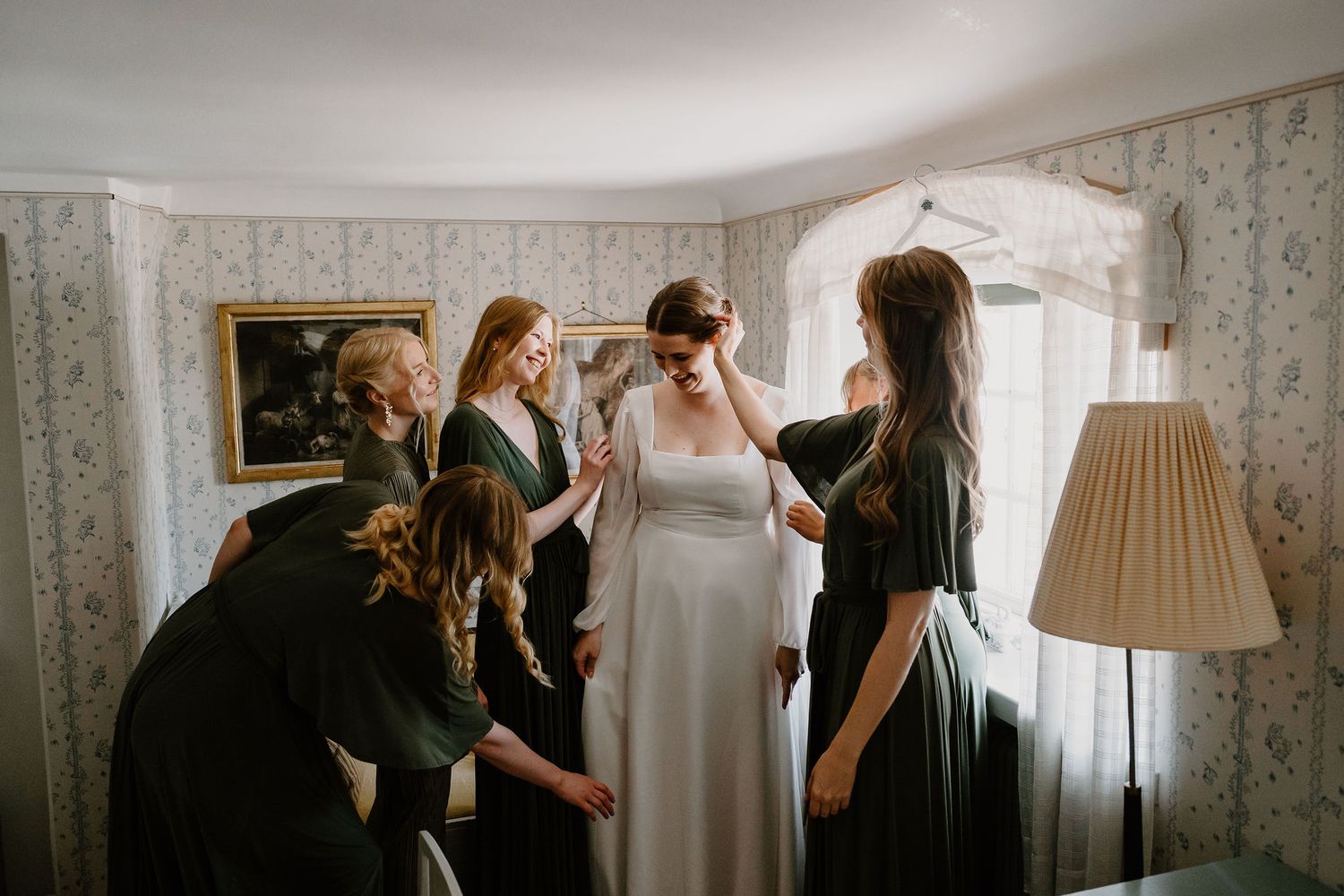 People gather in a room with floral wallpaper and window light as a bride prepares for her wedding day.