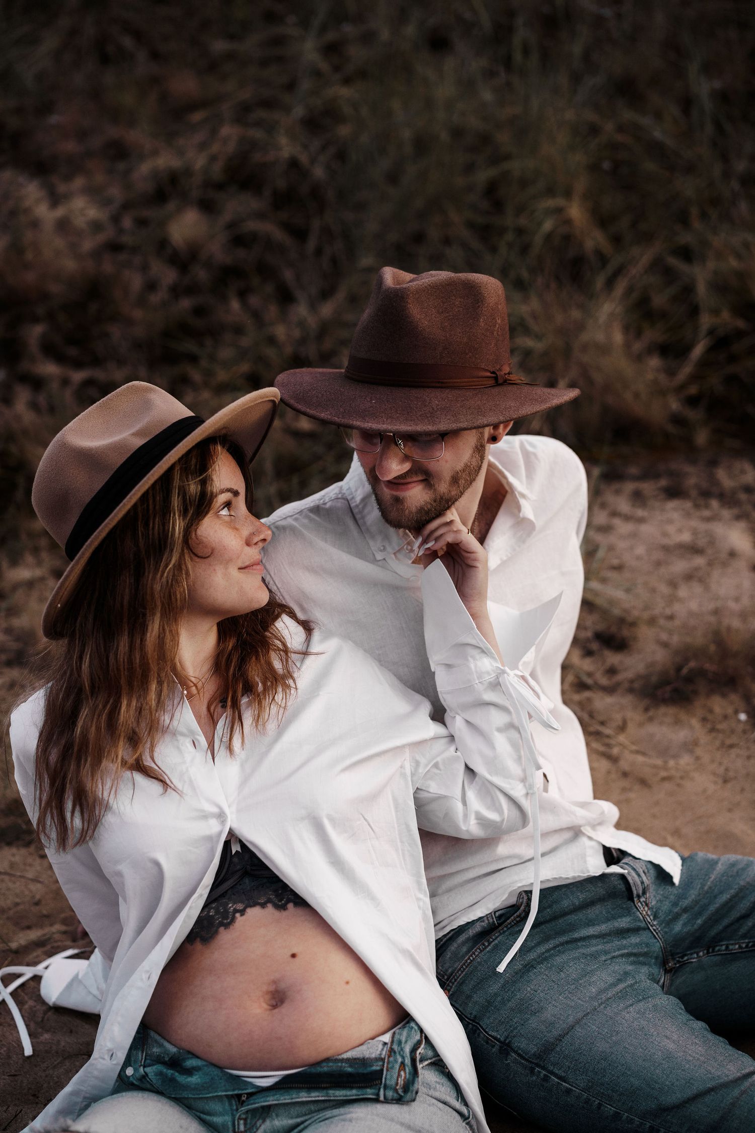Intimate maternity photo of couple wearing cowboy hats and white shirts against natural backdrop.