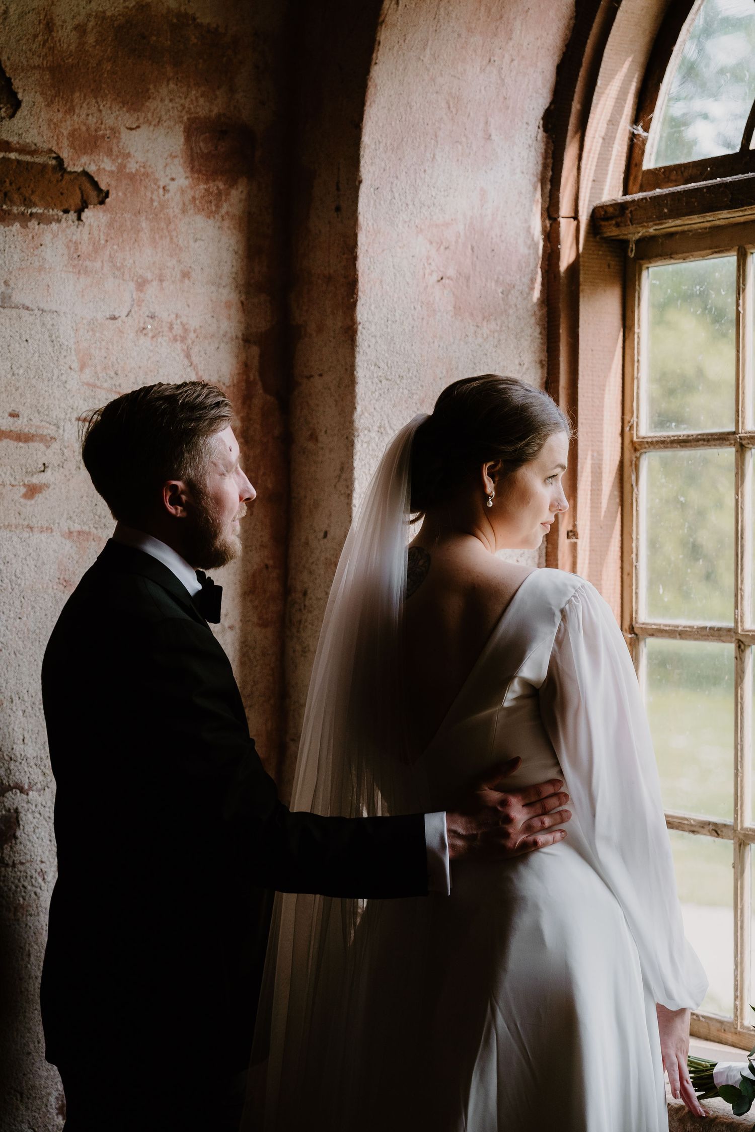 A couple stands by a rustic window in an intimate wedding moment, with soft natural light flowing in.