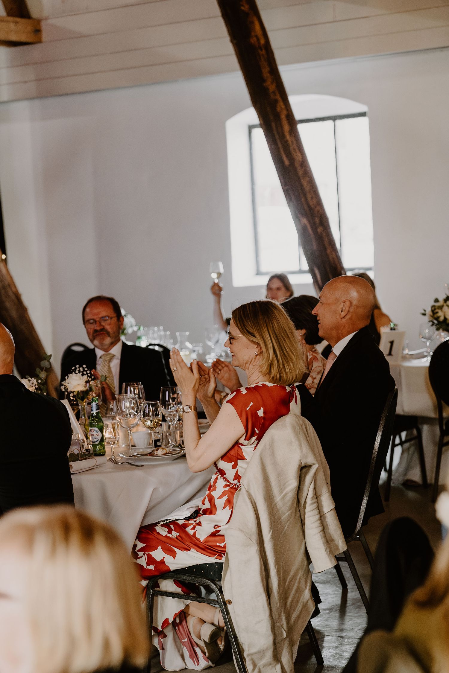 Wedding reception guests in formal attire dining at round tables in elegant venue with exposed wooden beams.