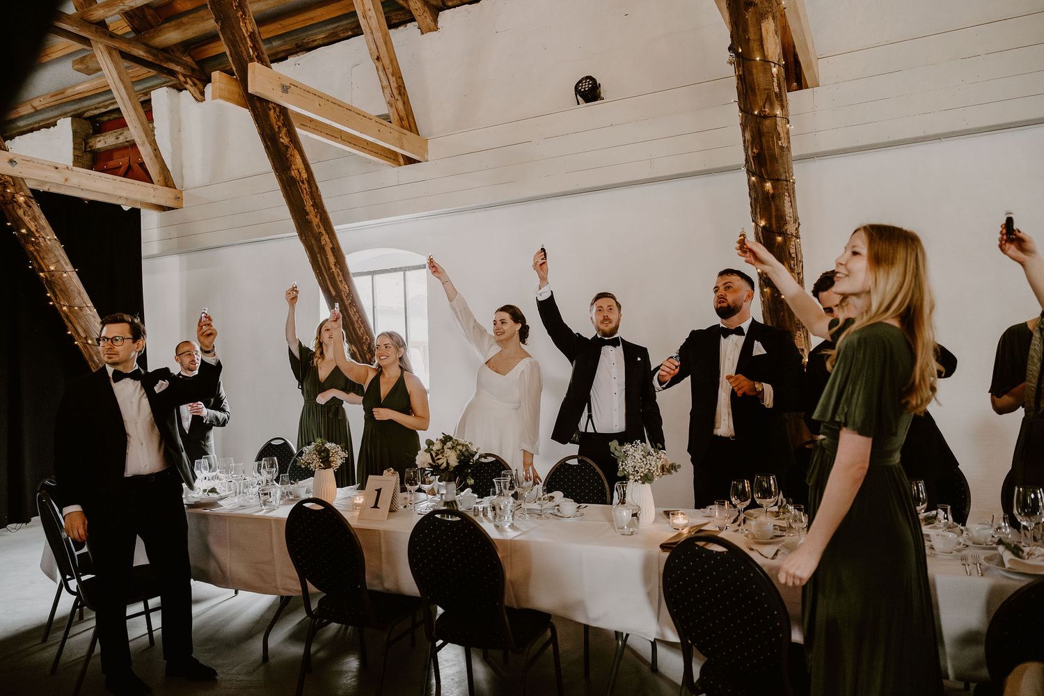 Wedding reception guests celebrate with raised hands and toasts at a long banquet table in a rustic venue.