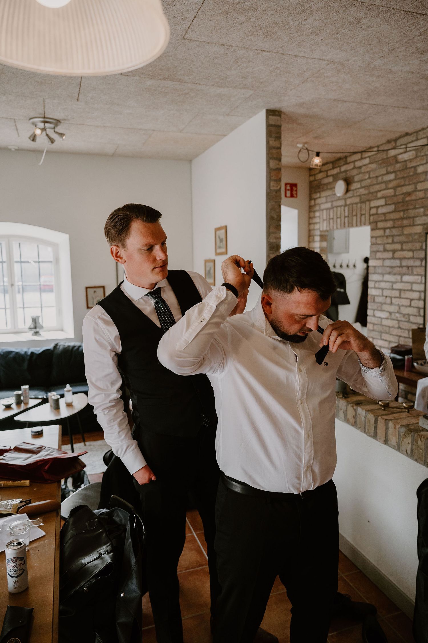 Two people in formal attire help each other get dressed in a modern room with exposed brick walls.