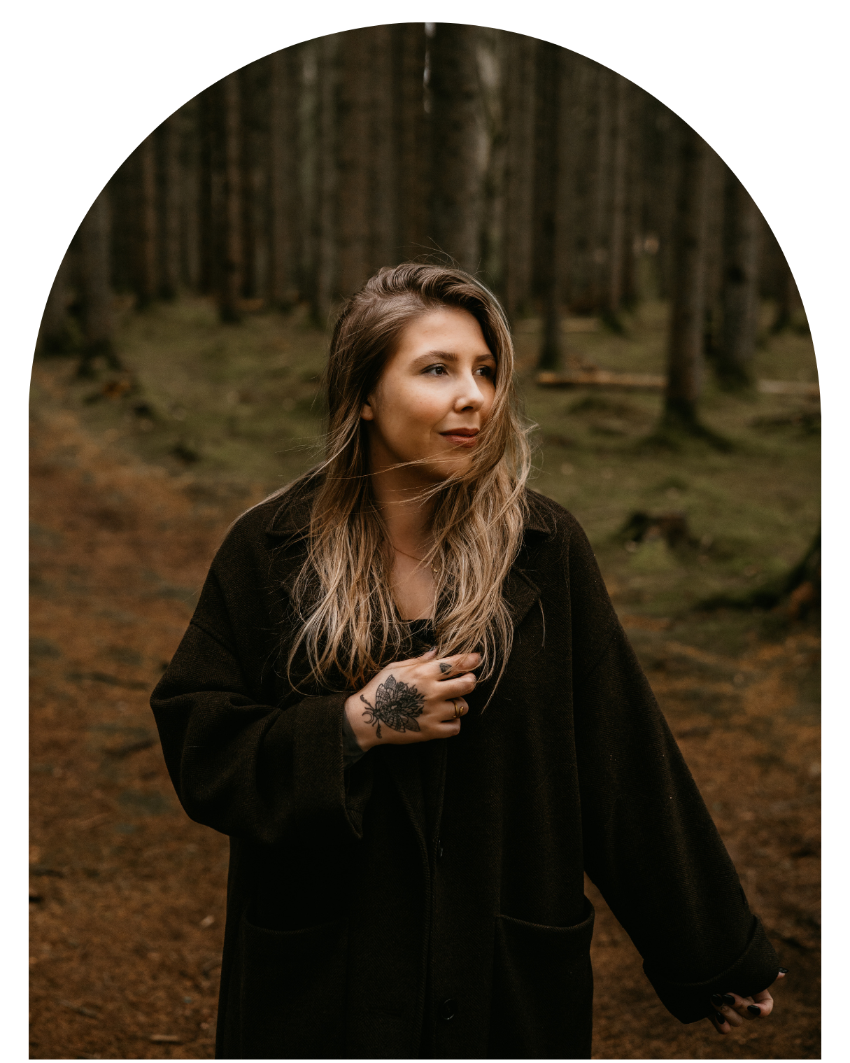 Person wearing a black winter coat poses thoughtfully in a moody forest setting with tall trees in the background.