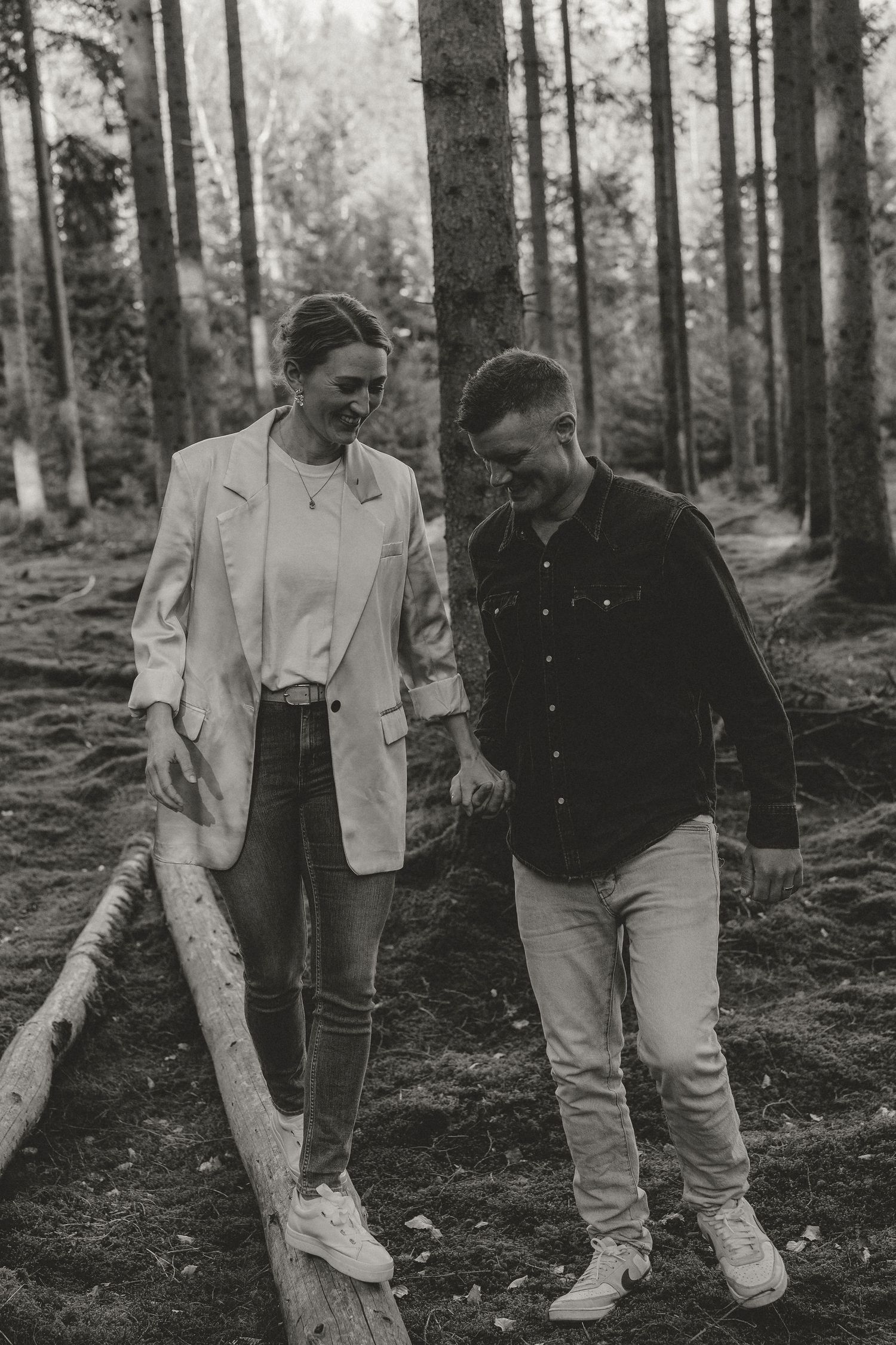 A couple walks together on a forest trail, wearing casual attire and sharing a candid moment in black and white.