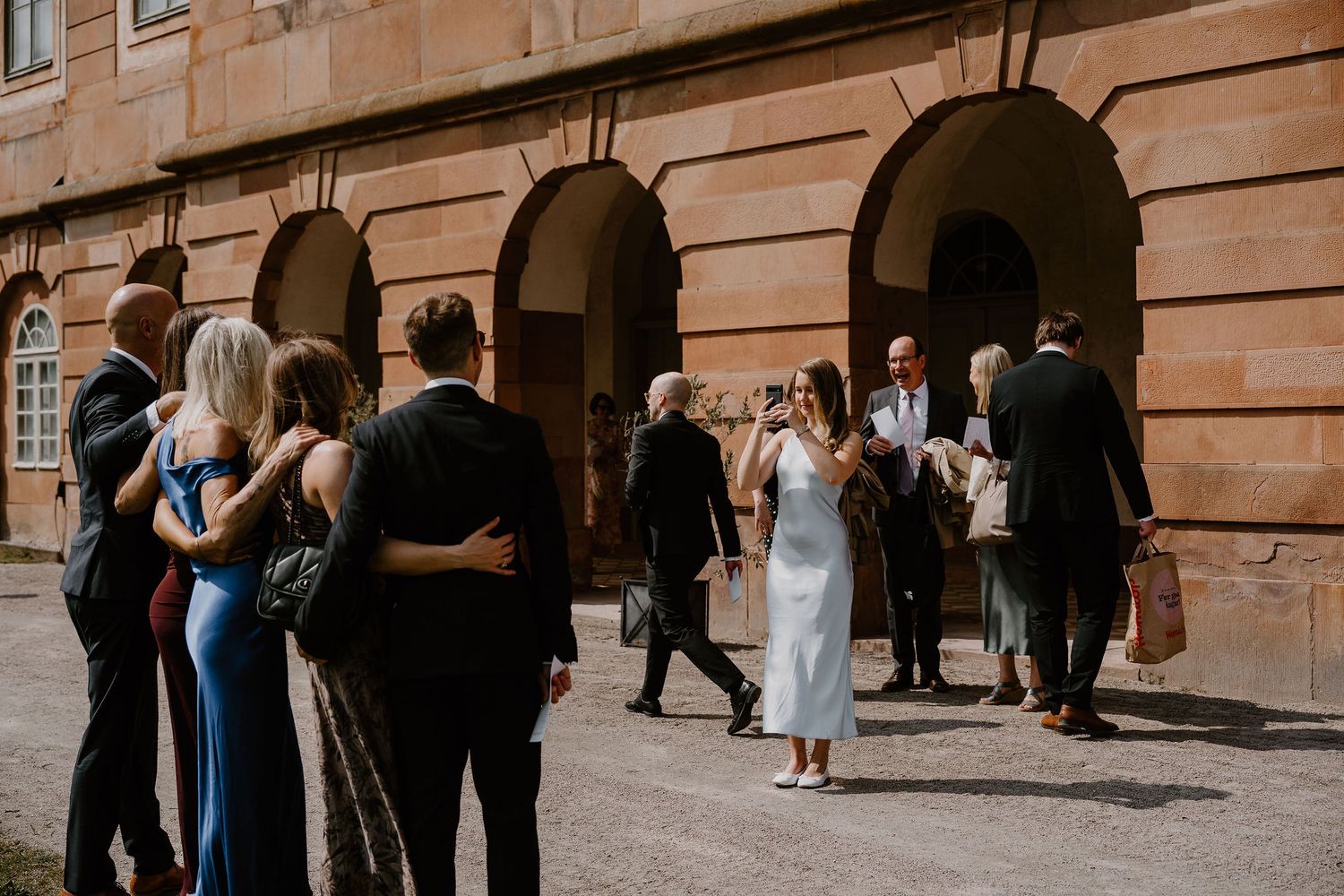 Wedding guests gather outside a stone building with archways during a formal celebration.