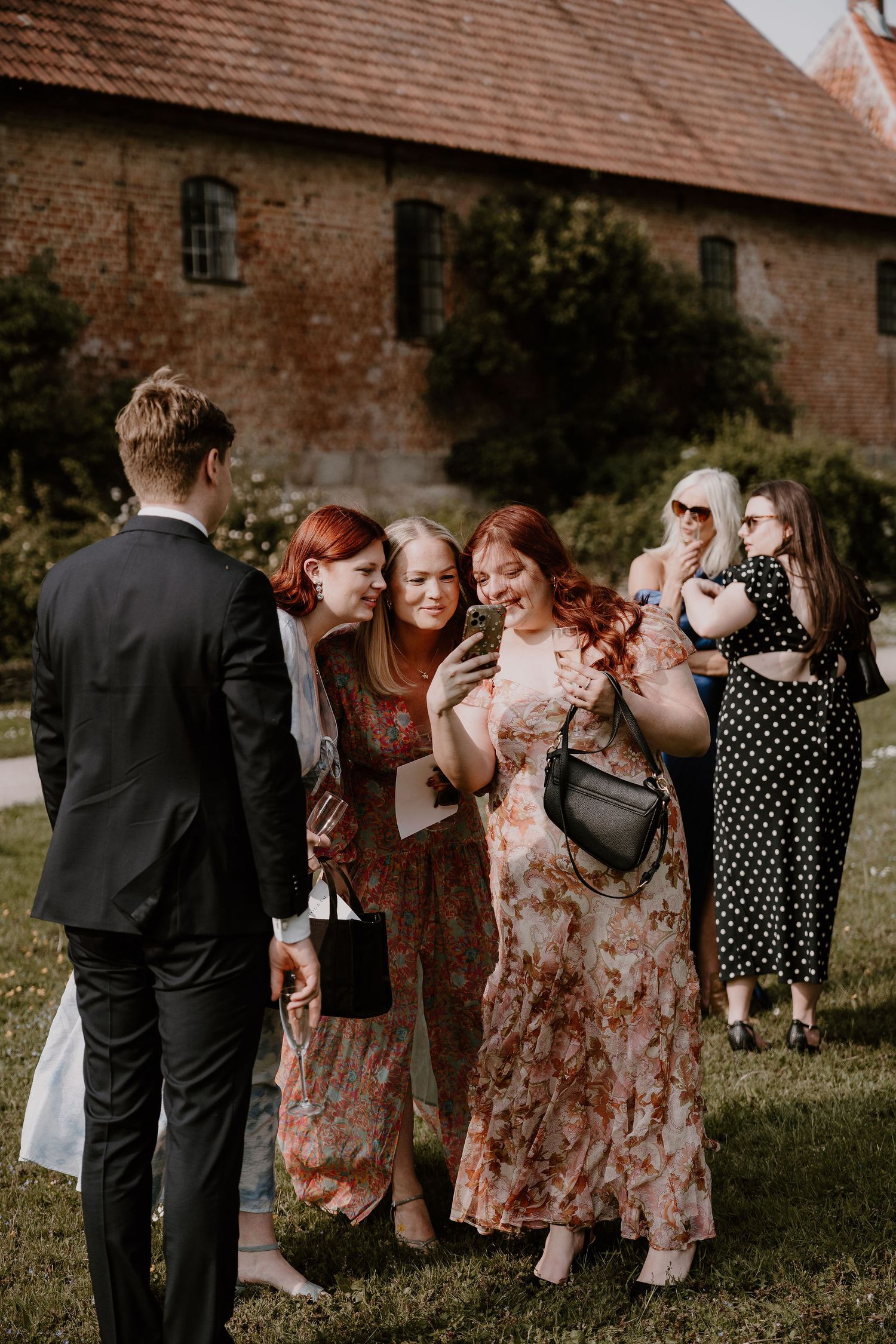 A wedding party in floral dresses and suits pose together while sharing laughter in a rustic outdoor setting.