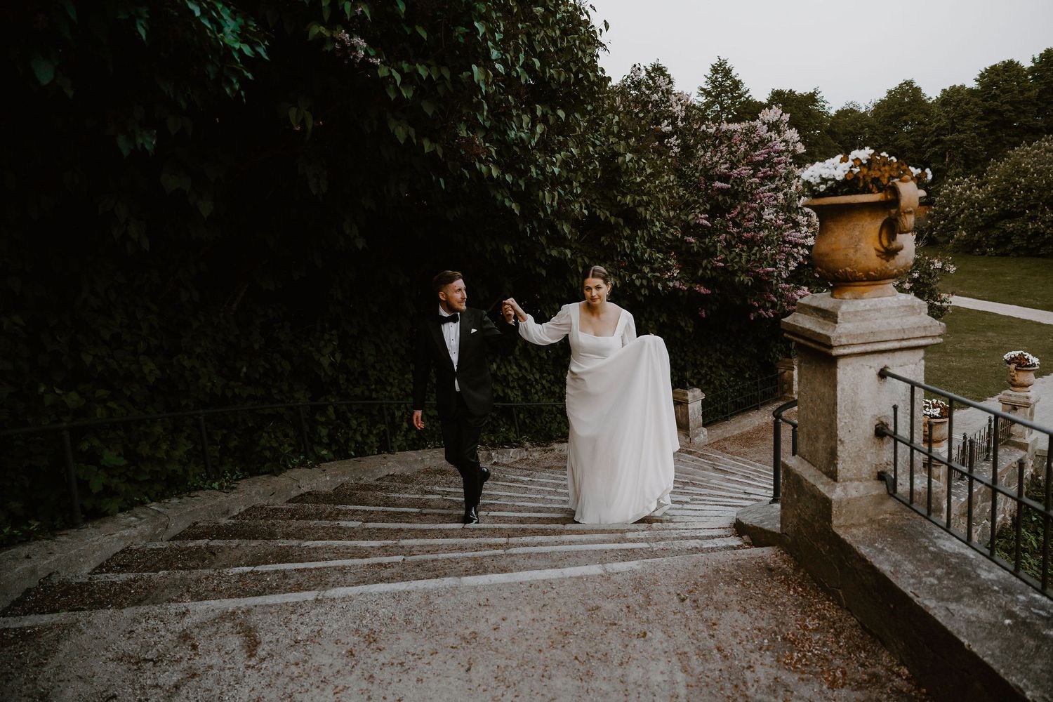 A couple in formal wedding attire walk up stone stairs in a garden setting at dusk.
