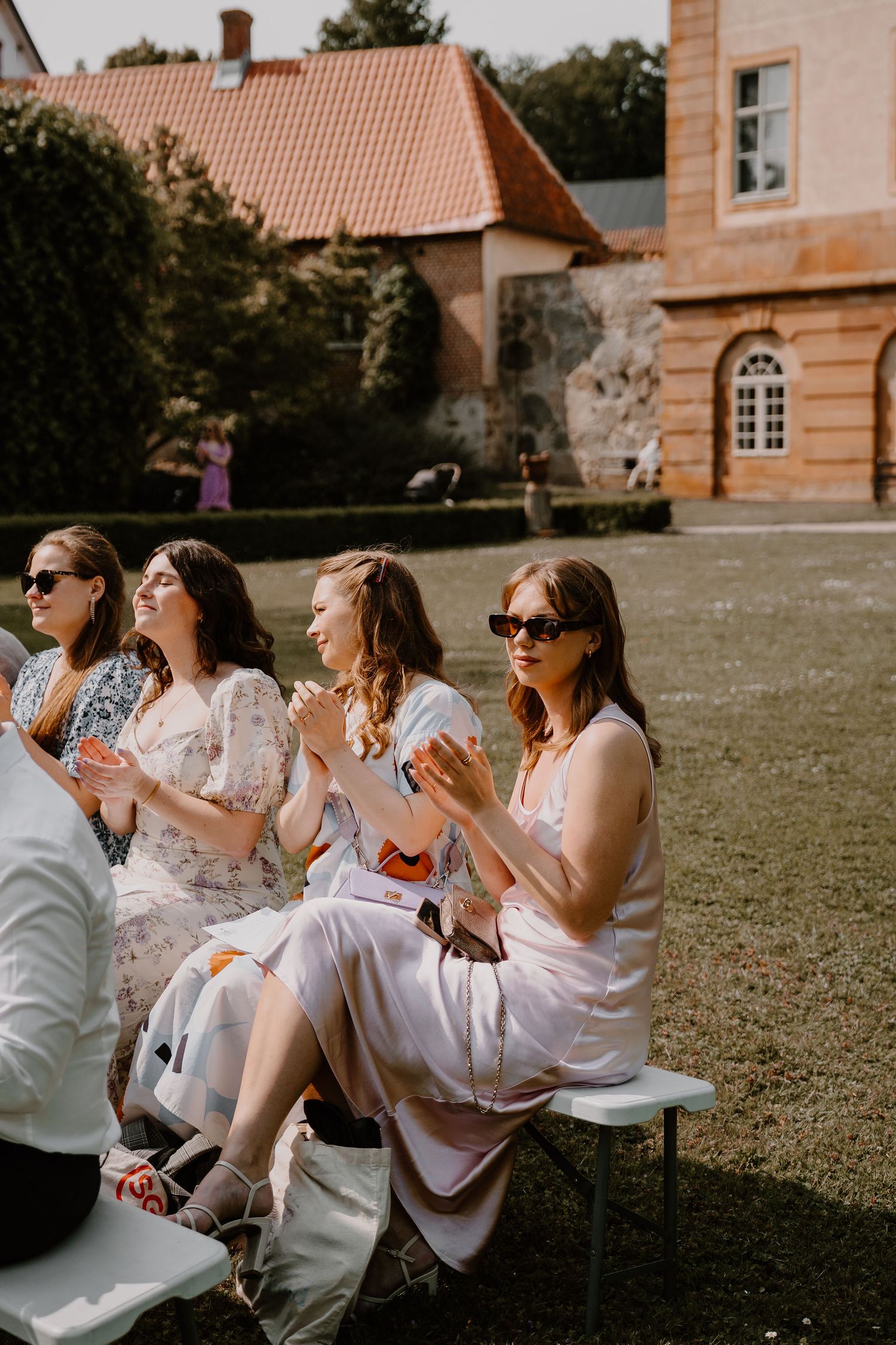 Wedding guests in summery dresses sit on outdoor benches enjoying refreshments at a beautiful estate venue.
