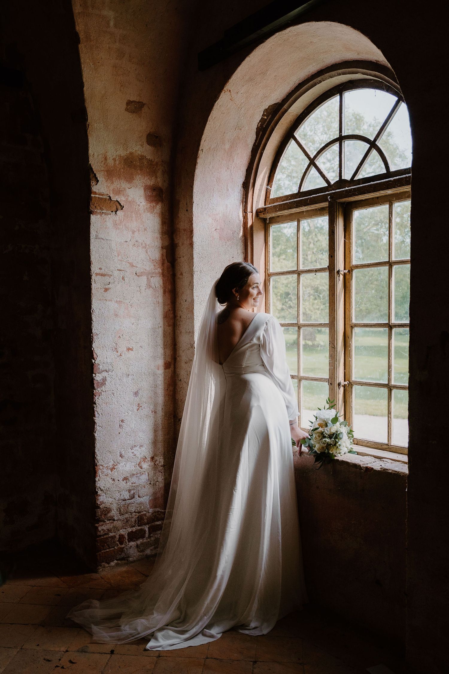 A bride in a flowing white gown stands elegantly by a large arched window in a rustic brick interior.