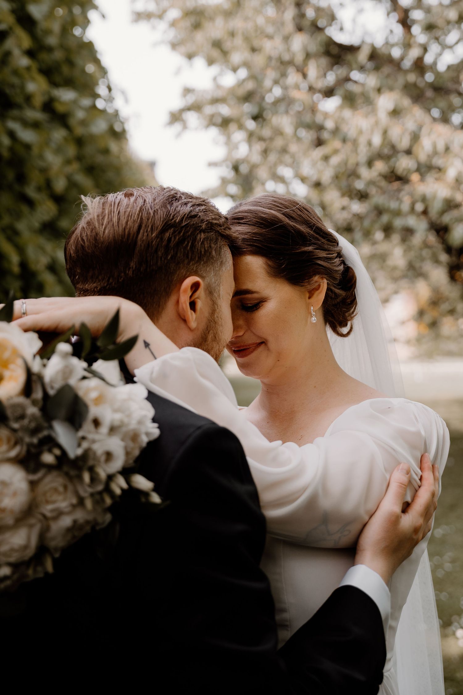 A romantic wedding couple embraces outdoors in soft light, with white flowers and elegant attire.