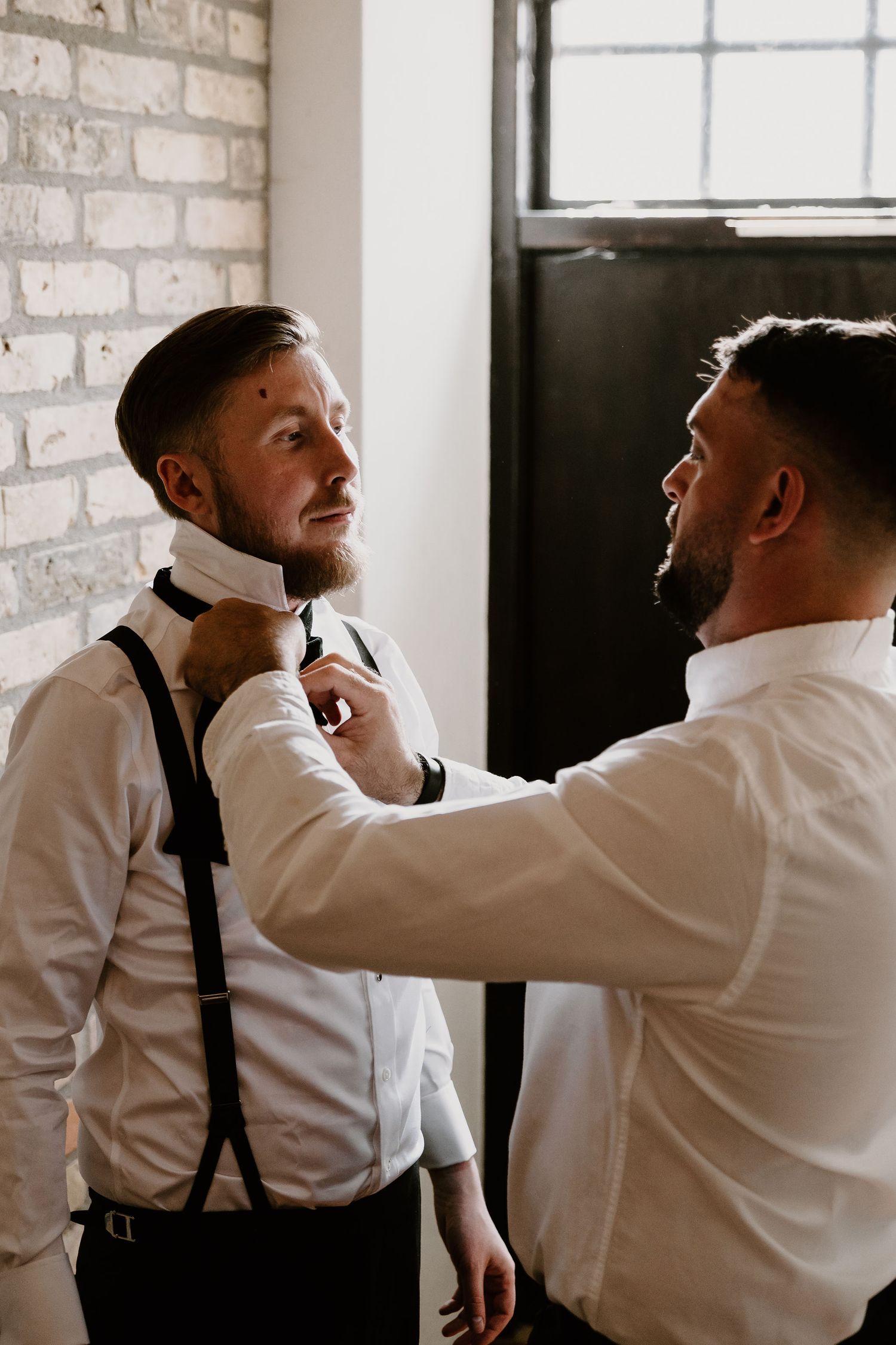 Two people in white shirts and suspenders help adjust a bowtie near a white brick wall and window.