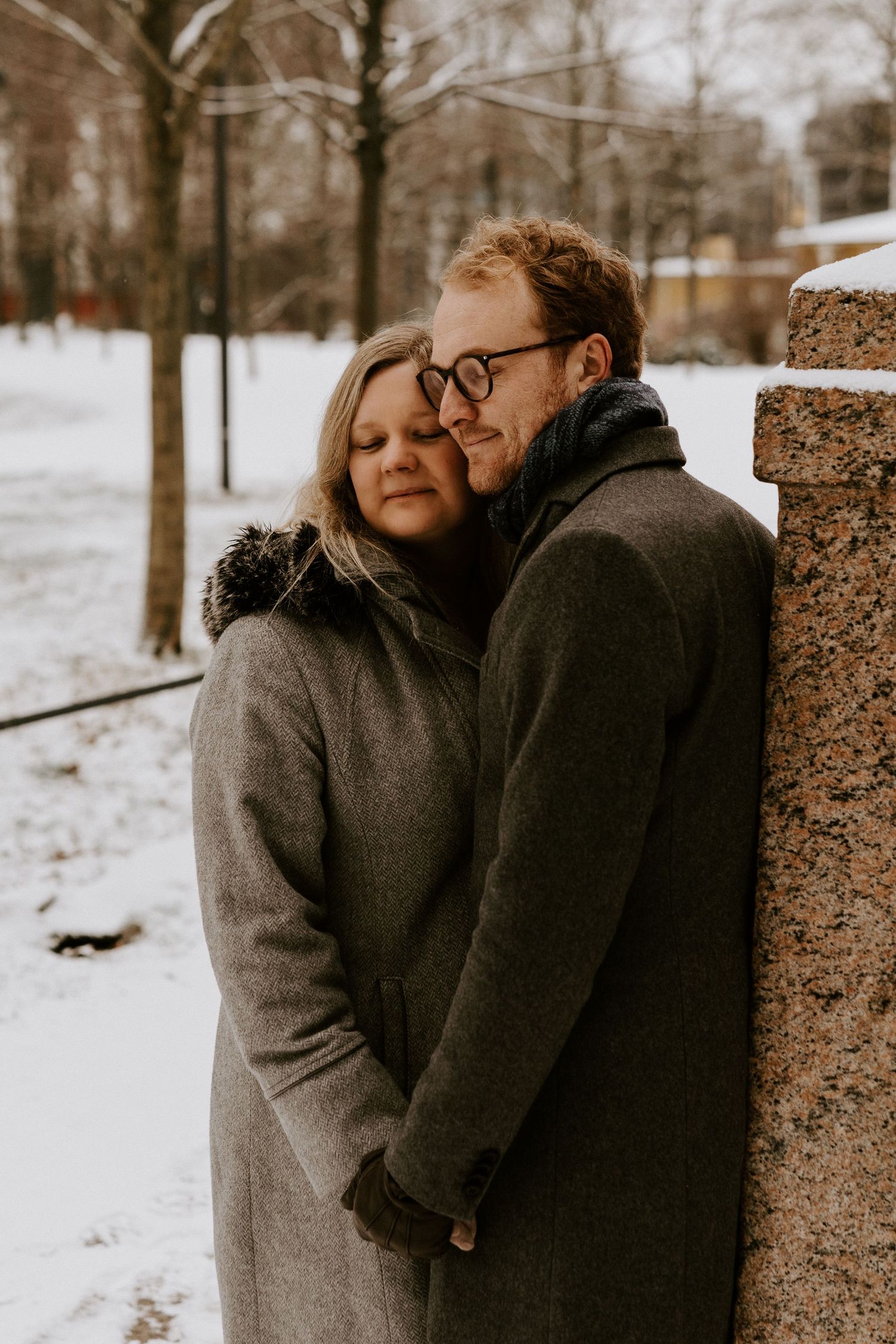 A couple in warm winter coats embrace against a brick wall with snowy trees in the background.