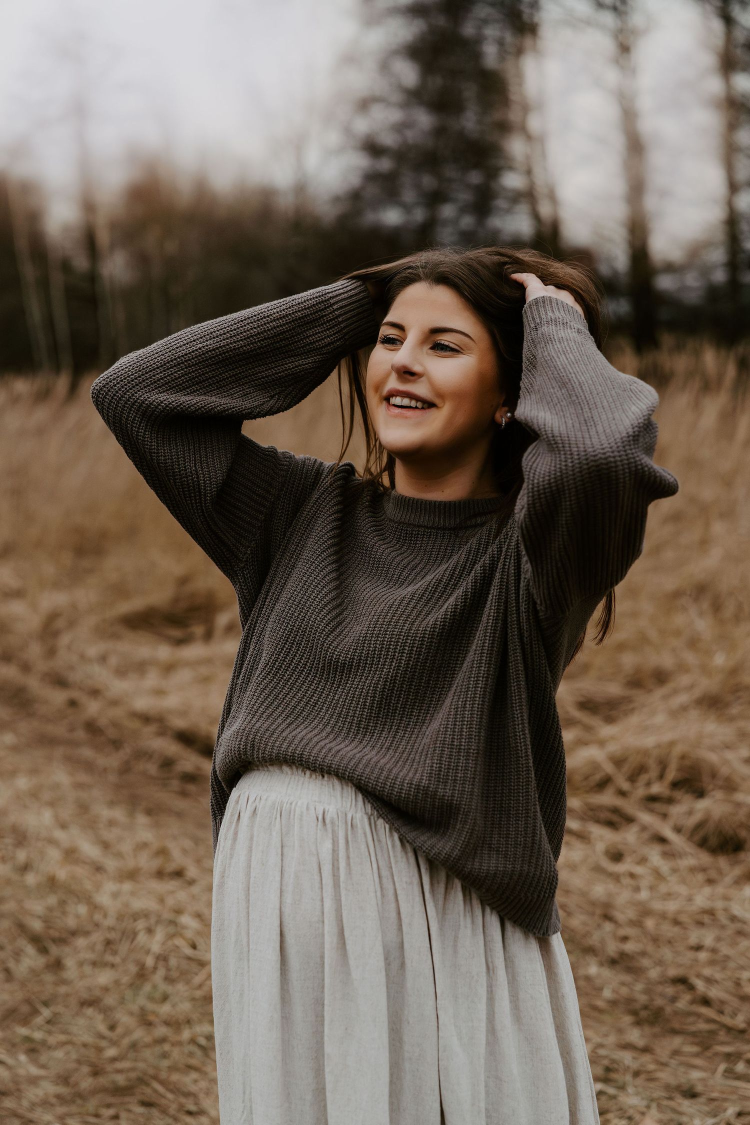 Person in oversized gray sweater and white skirt poses joyfully in a rustic outdoor setting at dusk.