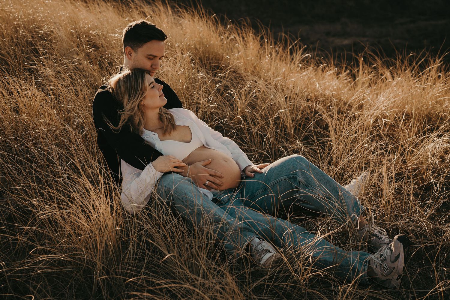 A couple shares a tender moment while sitting in golden grass on a hillside at sunset.