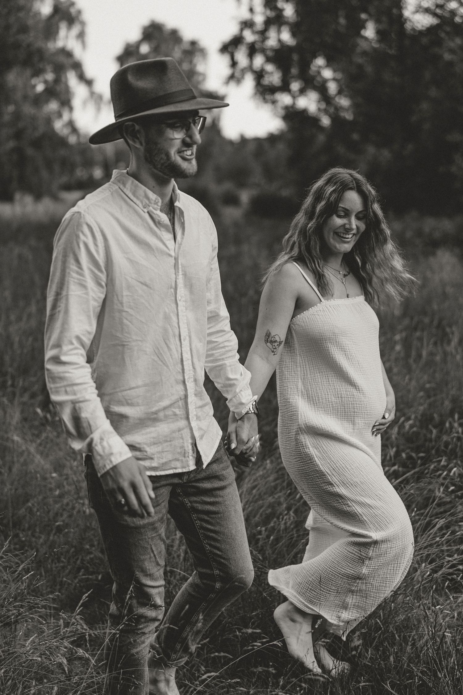A couple walks through a meadow holding hands, with her wearing a white dress and him in a hat and button-down shirt.
