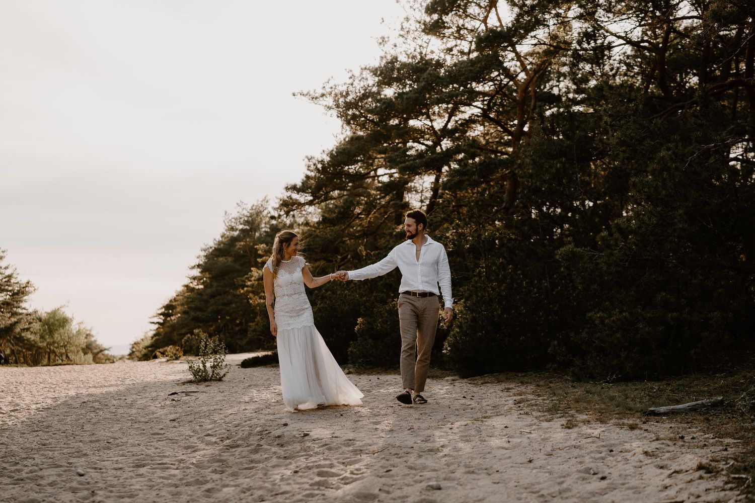 A couple in wedding attire holds hands while walking on a sandy beach path with tall pine trees in the background at sunset.