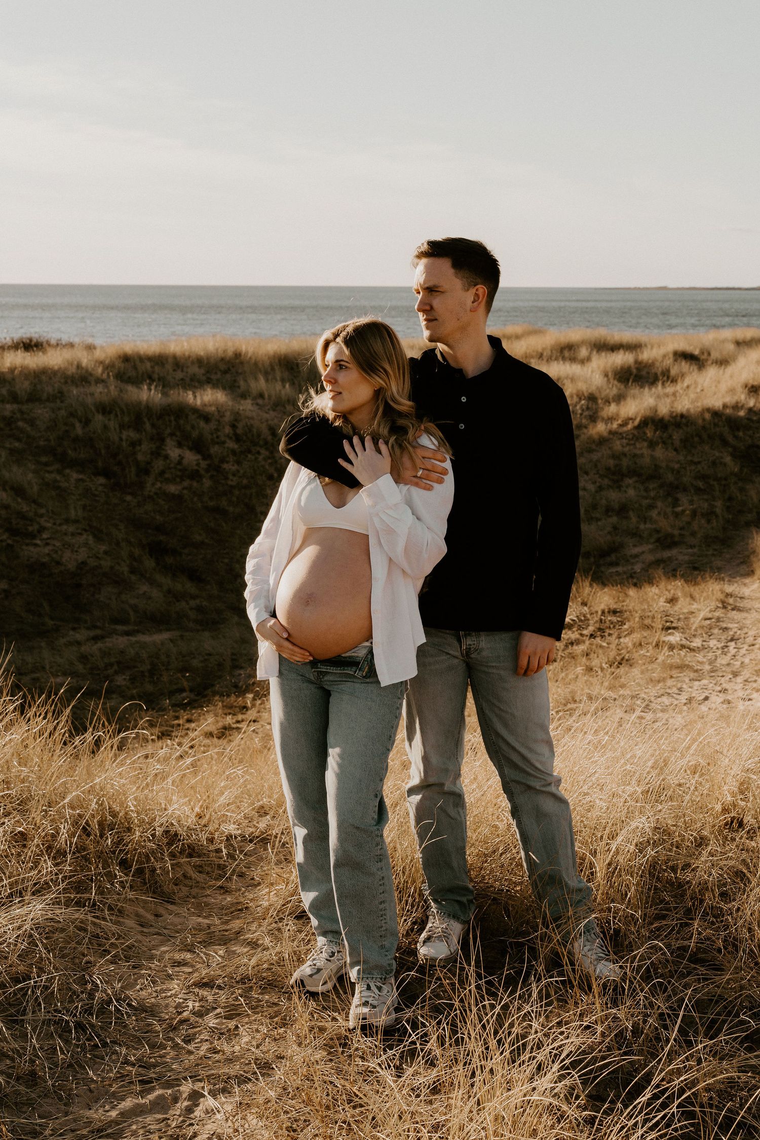 Expecting couple embraces while standing on grassy coastal cliffside at sunset overlooking the ocean.