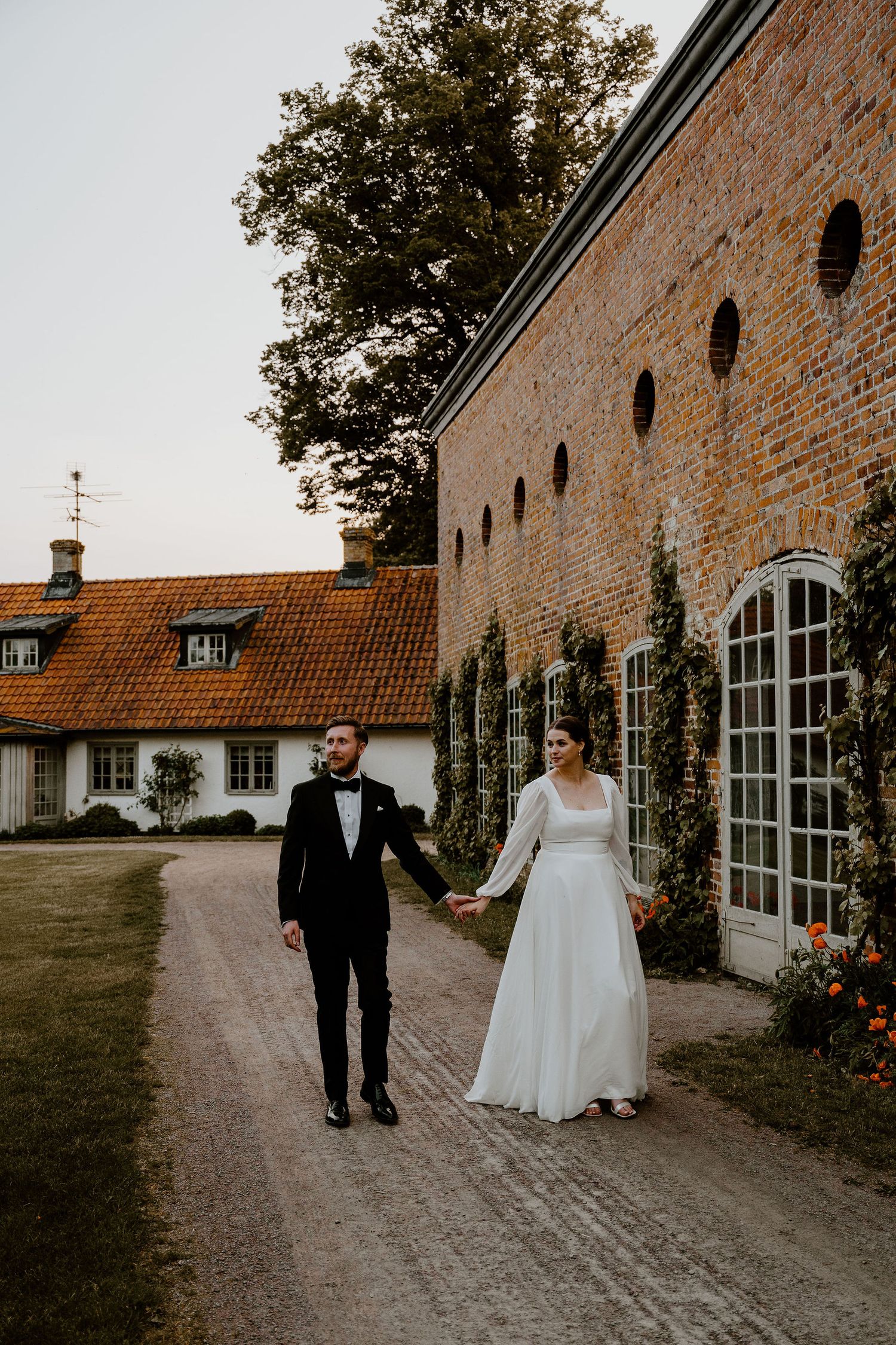 A couple walking together on a rustic outdoor pathway next to a vintage brick building.