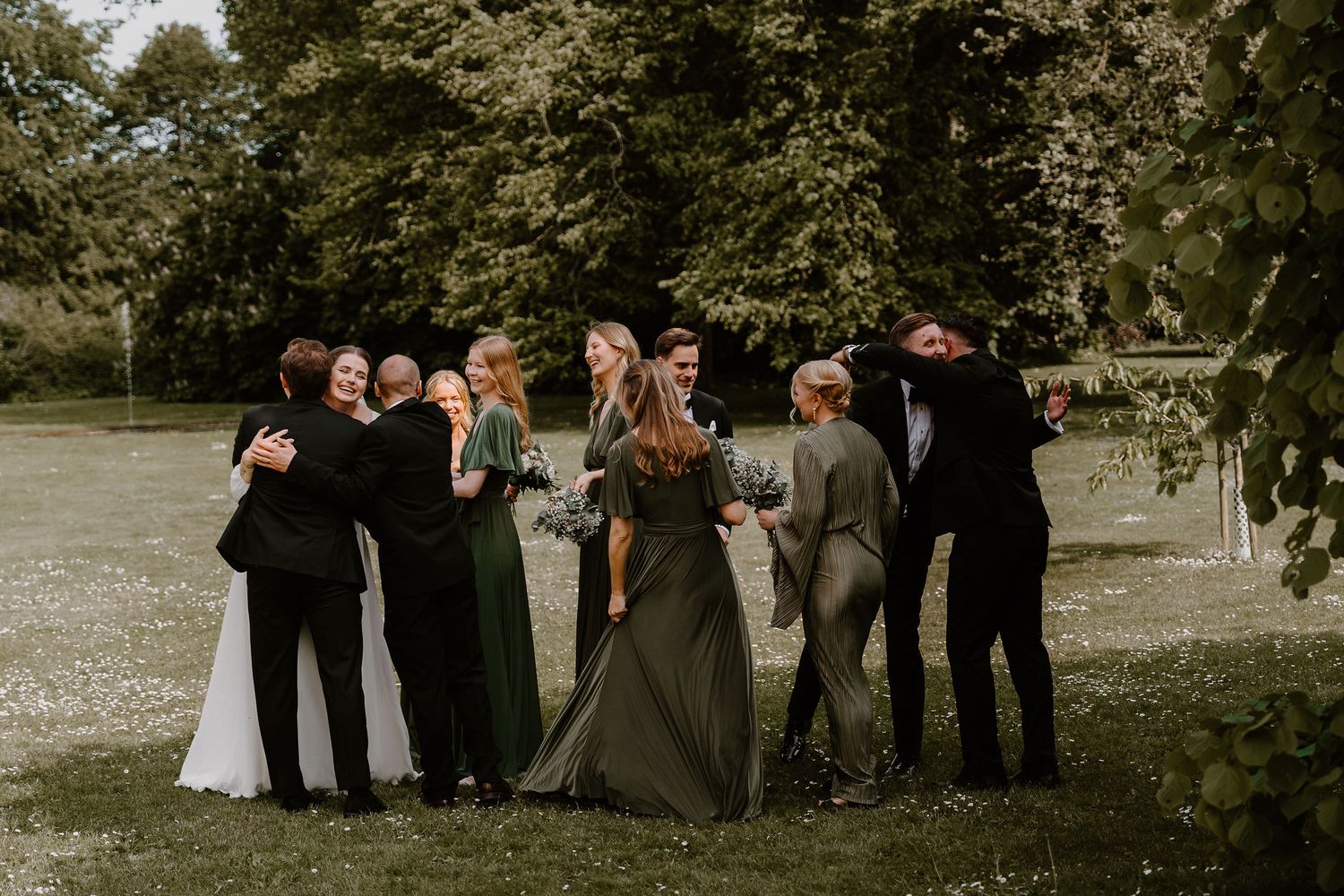 Wedding party gathers outdoors for group photos in a casual natural setting with trees in the background.