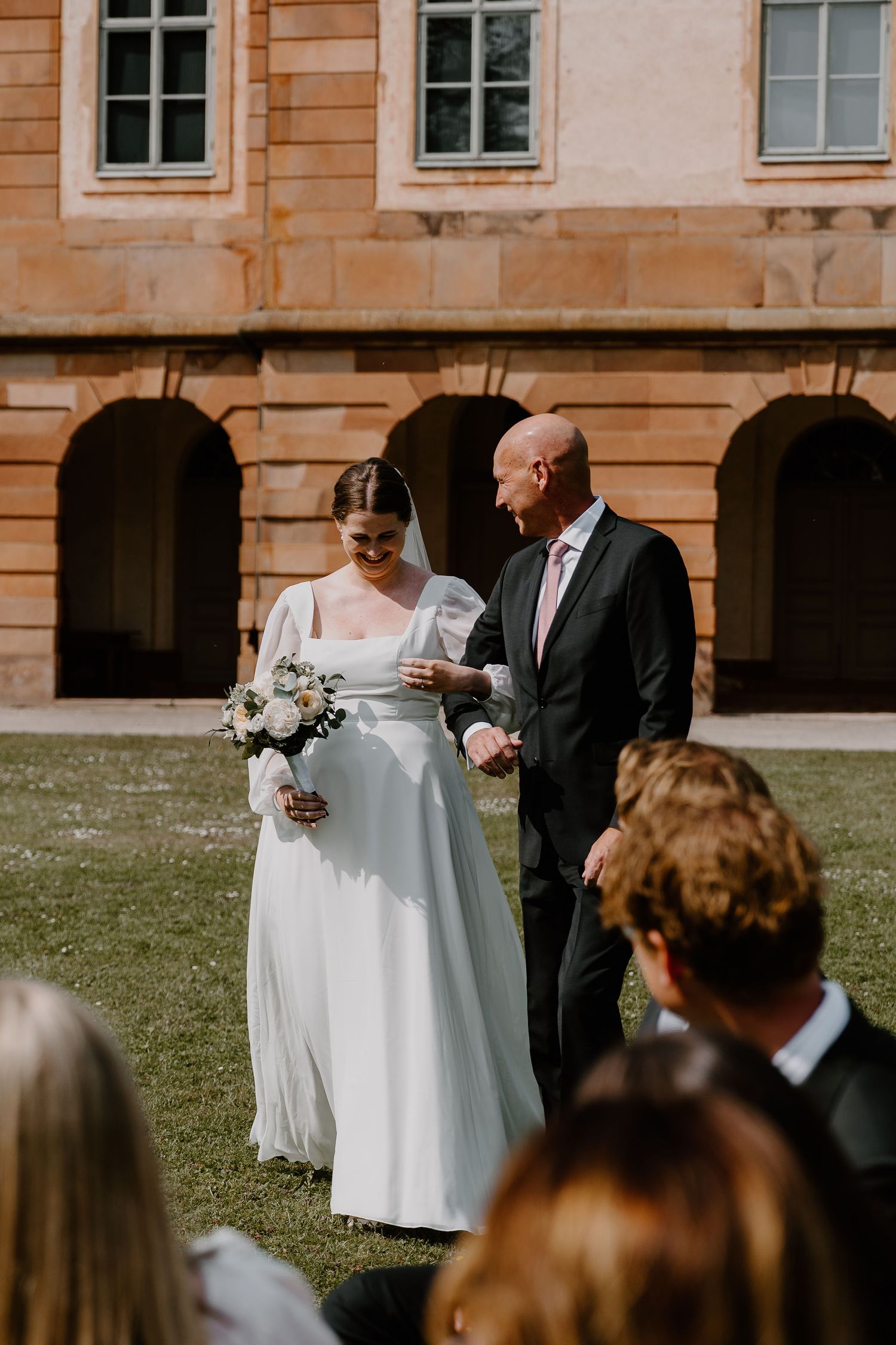 An intimate outdoor wedding ceremony takes place in front of a historic stone building.