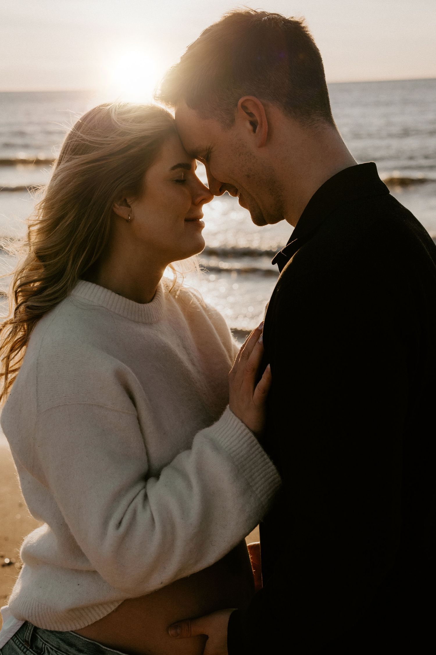 Romantic couple embraces on beach at sunset with golden light illuminating their silhouettes against ocean backdrop.