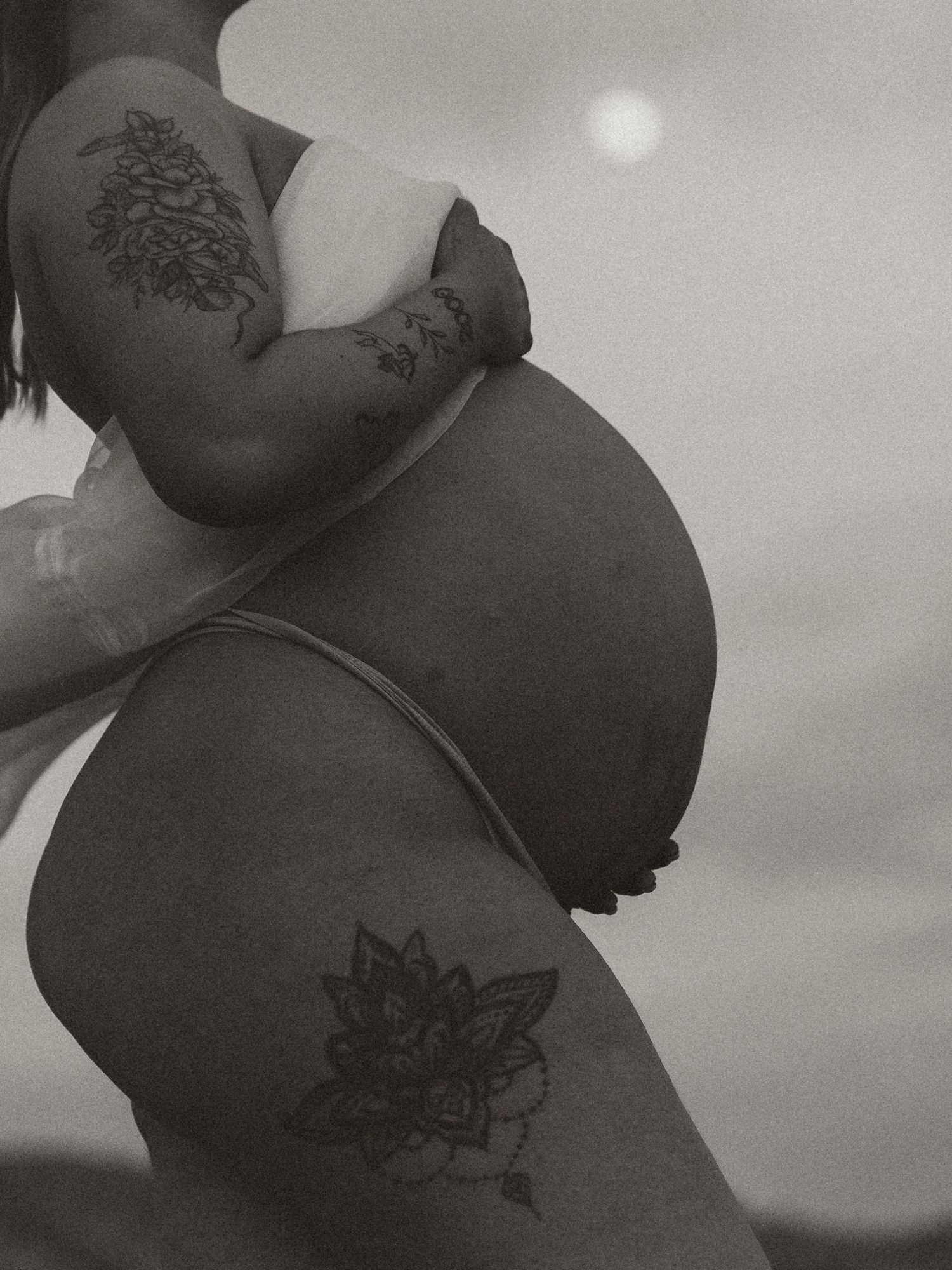 Black and white photograph showing two nature-inspired tattoos on a person's body against a hazy sky with visible sun.