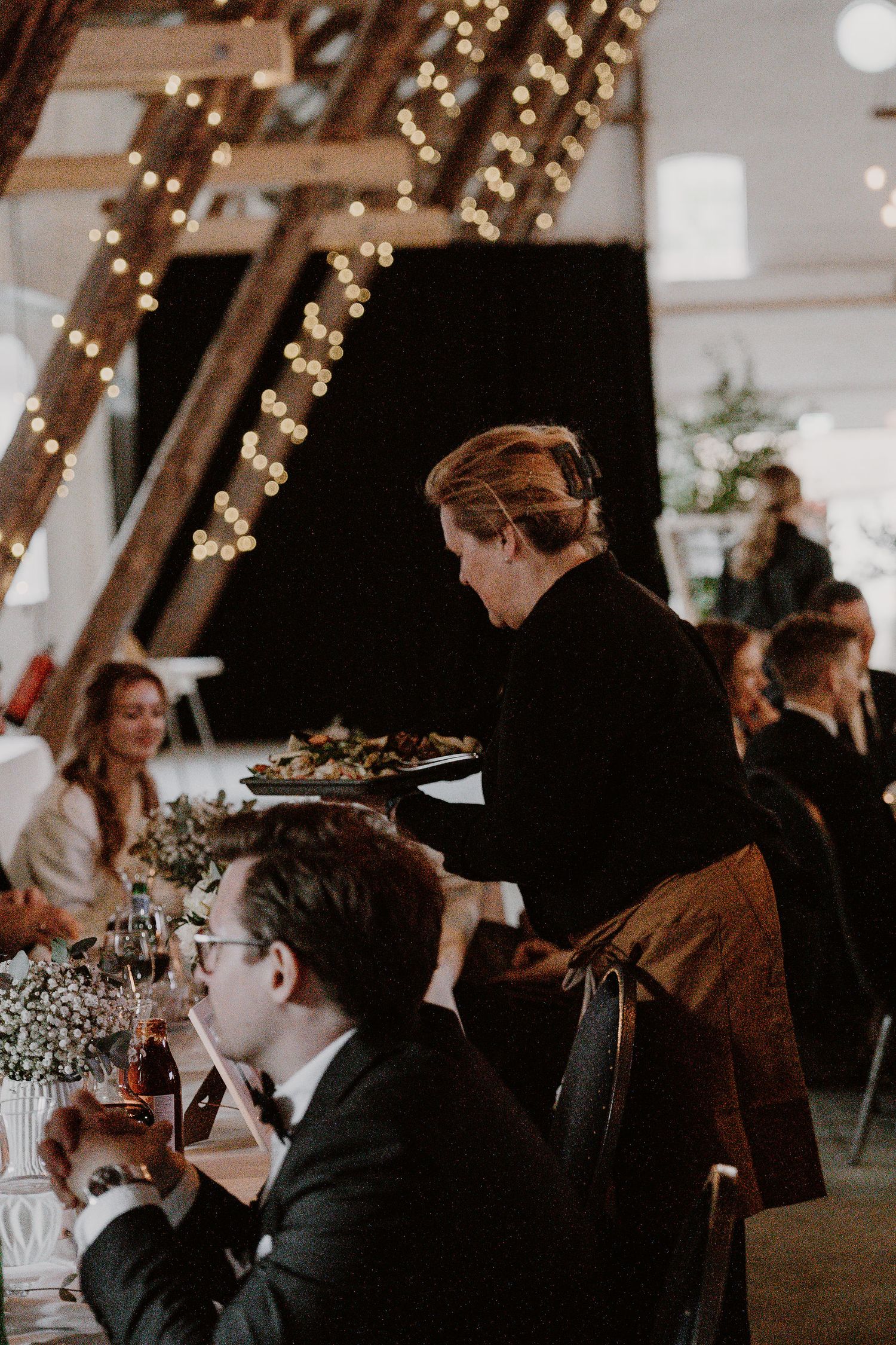 A server carries plates at an indoor wedding reception decorated with romantic string lights overhead.