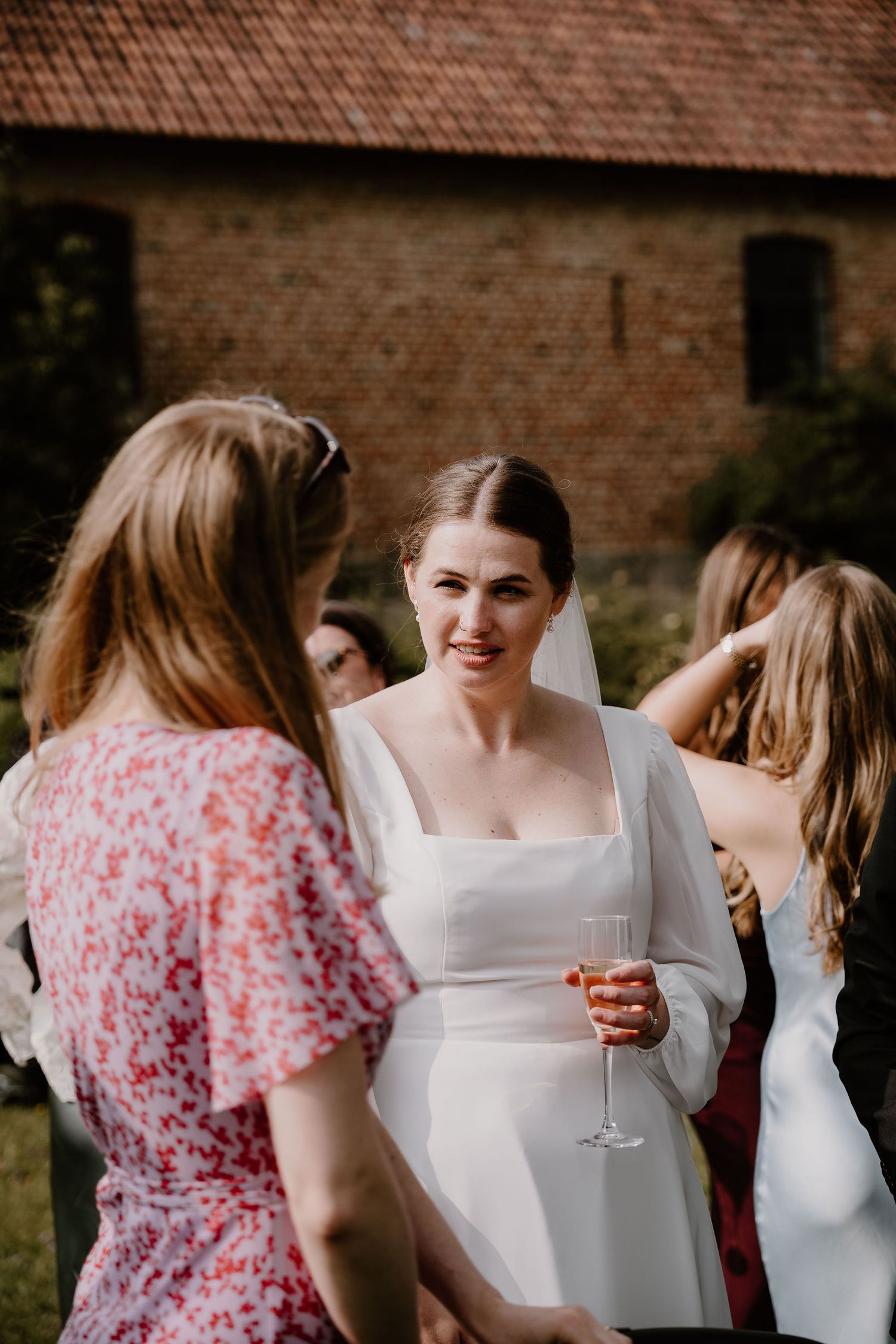 A bride in a modern white wedding dress holds a glass of wine while chatting with guests at an outdoor reception.