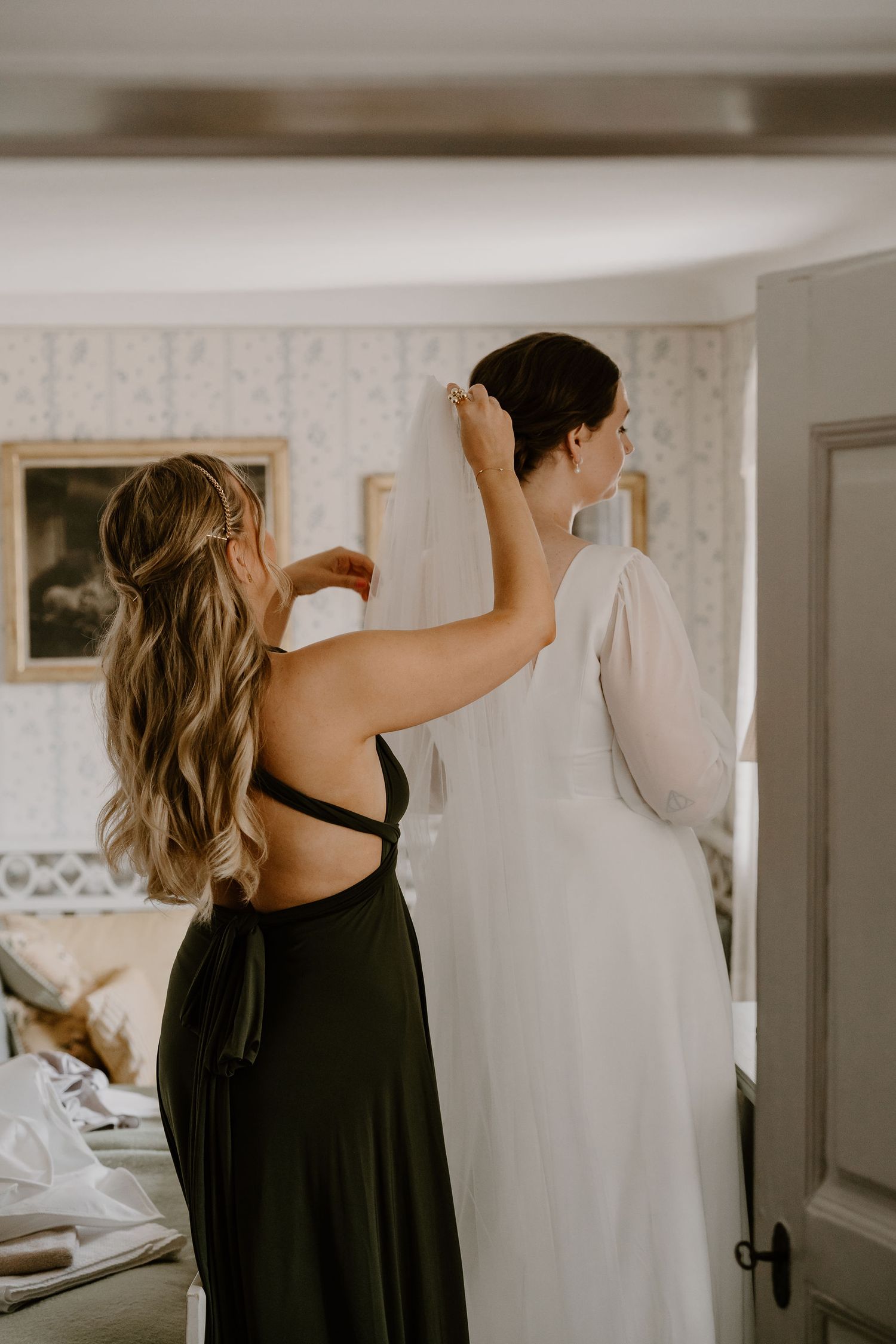 A bridesmaid in a black dress helps adjust a flowing white wedding gown in a sunlit dressing room.