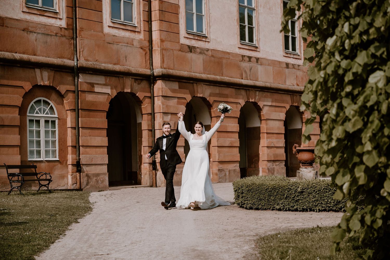 A couple in wedding attire poses outside a grand historic building with stone walls and elegant windows.